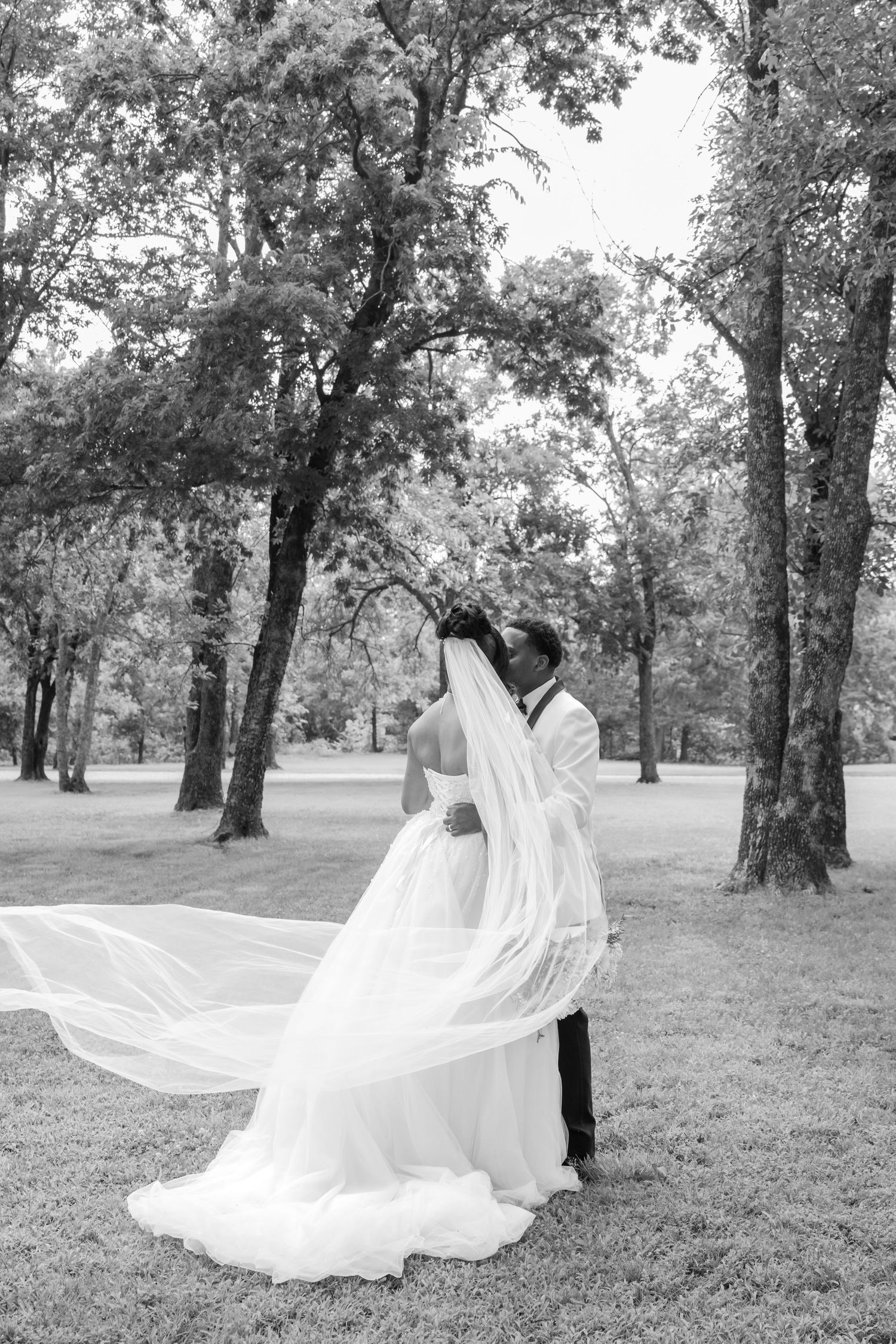 Black and white photo of bride and groom embracing outdoors, veil flowing in the wind among tall trees.