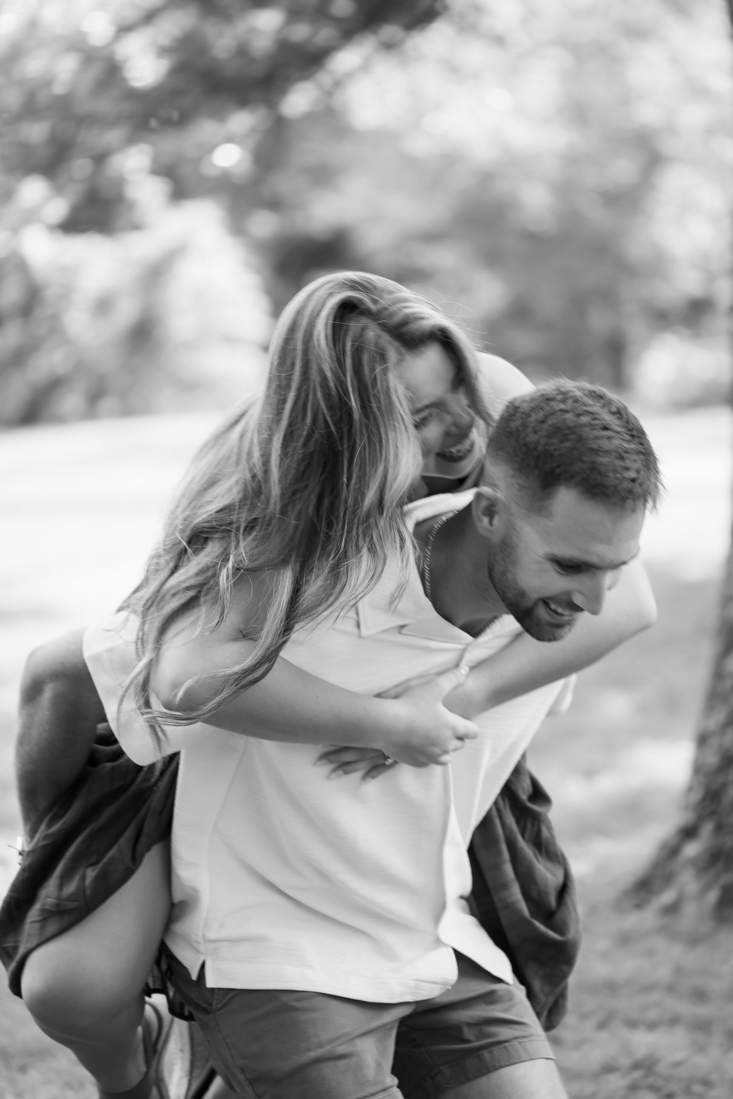 A black and white photo of a man giving a laughing woman a piggyback ride in a park.