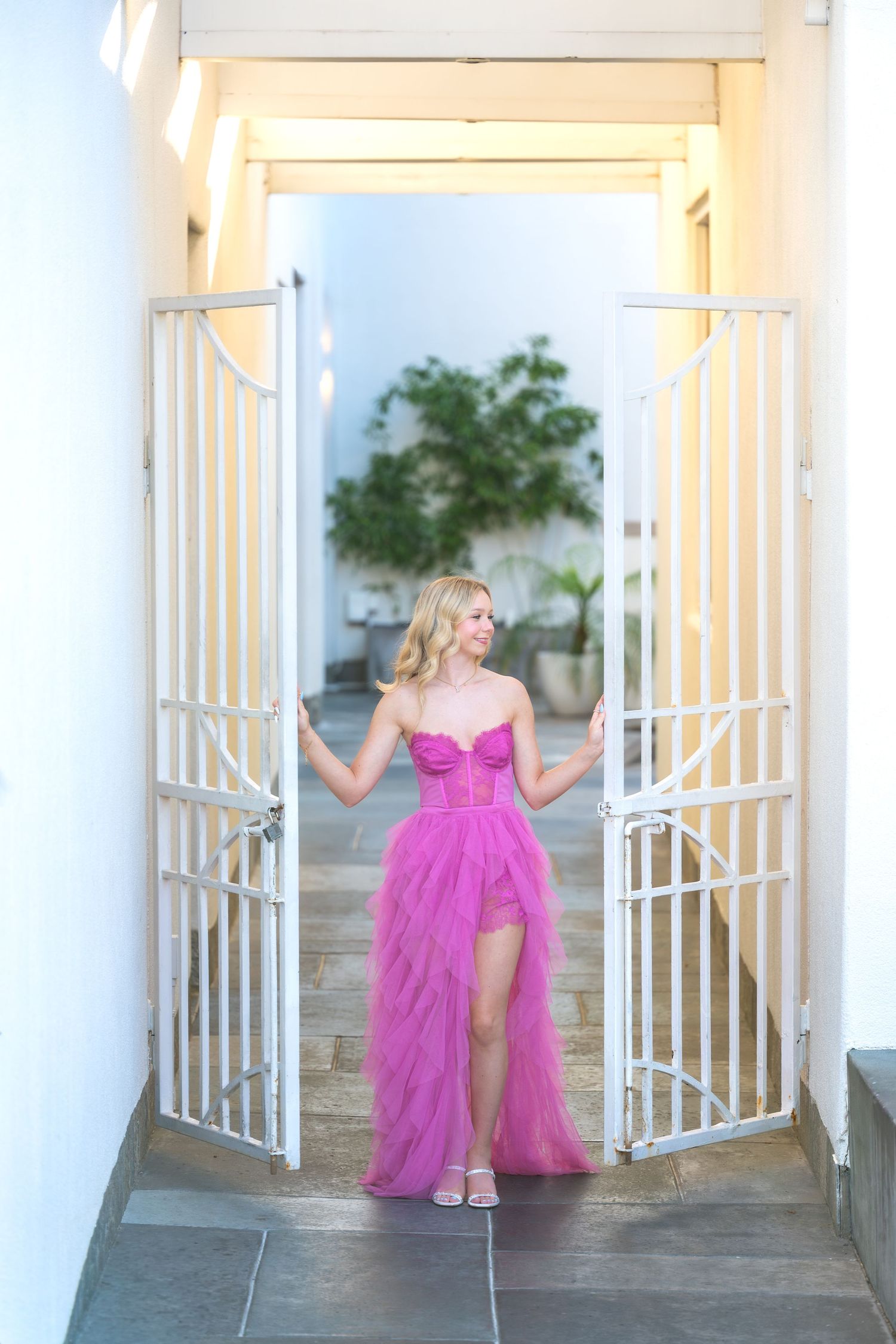 Blonde woman in a strapless pink tulle gown standing in white iron gate doorway.