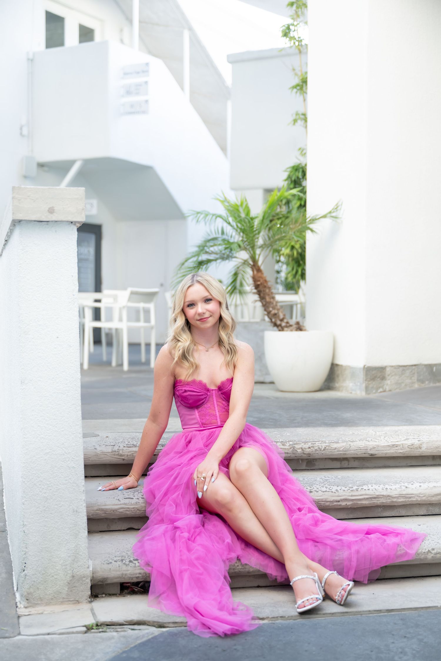Blonde woman in a pink tulle gown sitting on steps outdoors near a white building and palm tree.