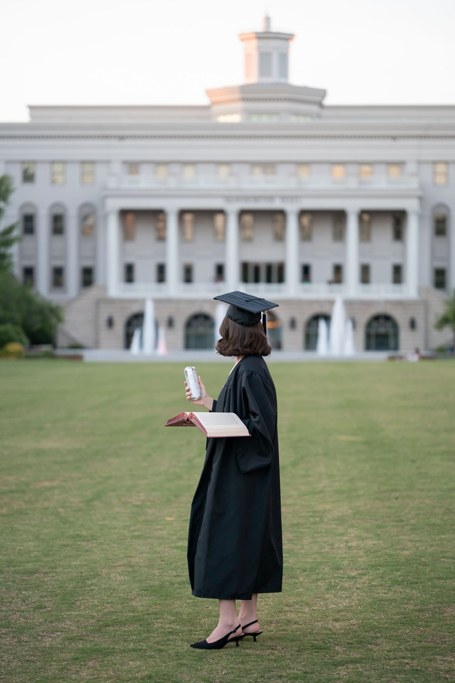 Female graduate in black cap and gown holds diploma and book on college campus lawn.