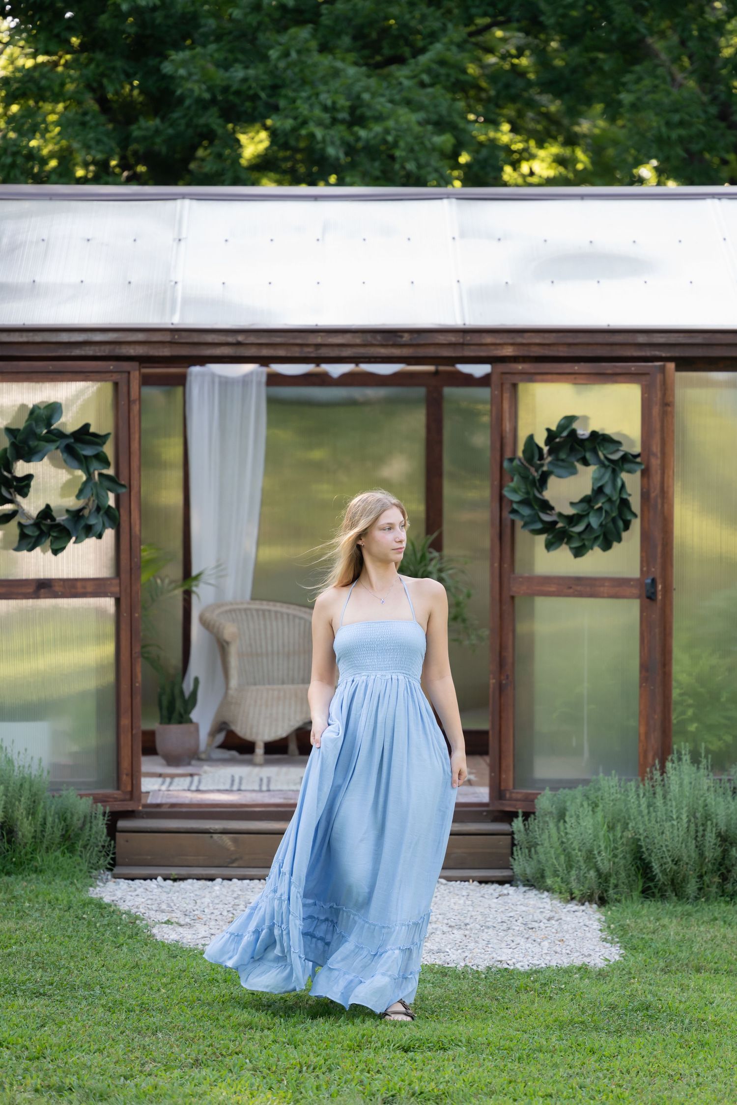 Young woman in flowing blue strapless maxi dress standing on green lawn in front of glass greenhouse with wreaths.