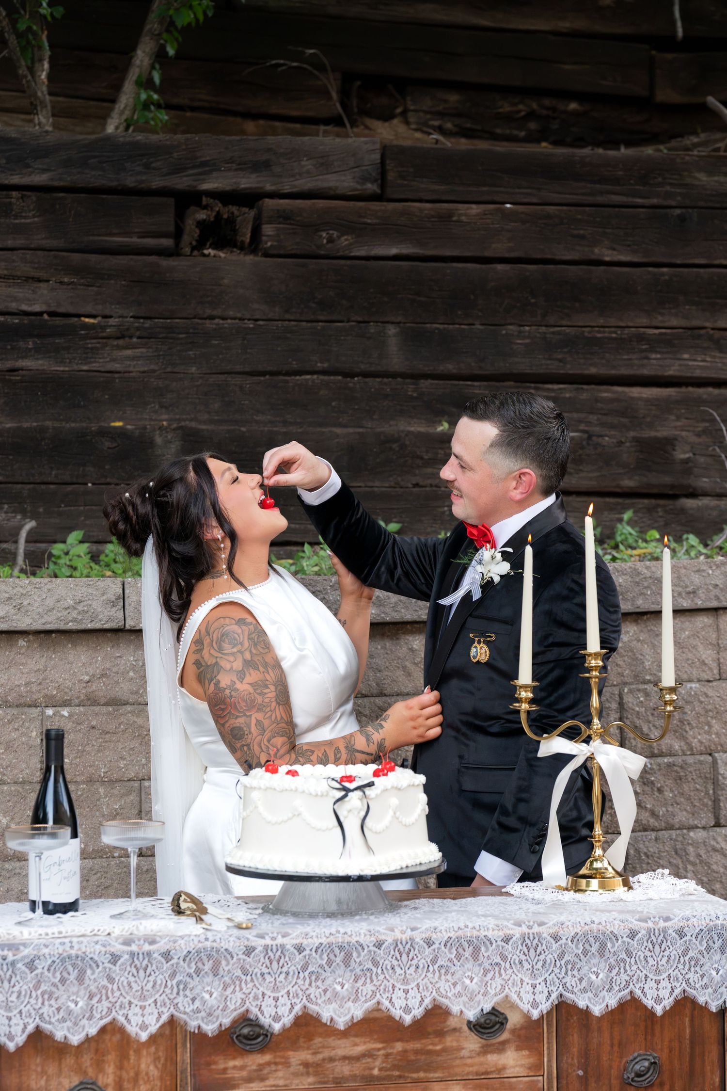 Bride and groom cutting wedding cake outdoors, groom feeding bride near candlesticks and wine glasses on lace-covered table.