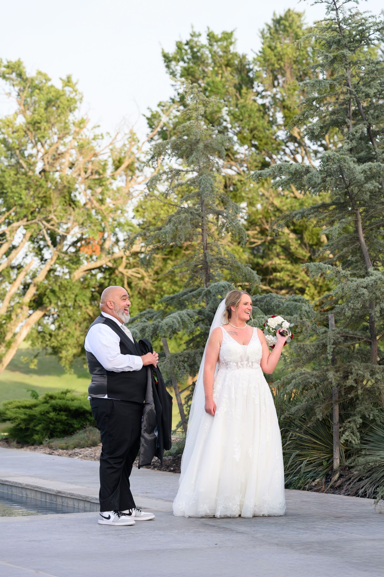 Bride in white gown holding bouquet shares first look moment with groom in vest outdoors near lush green trees.