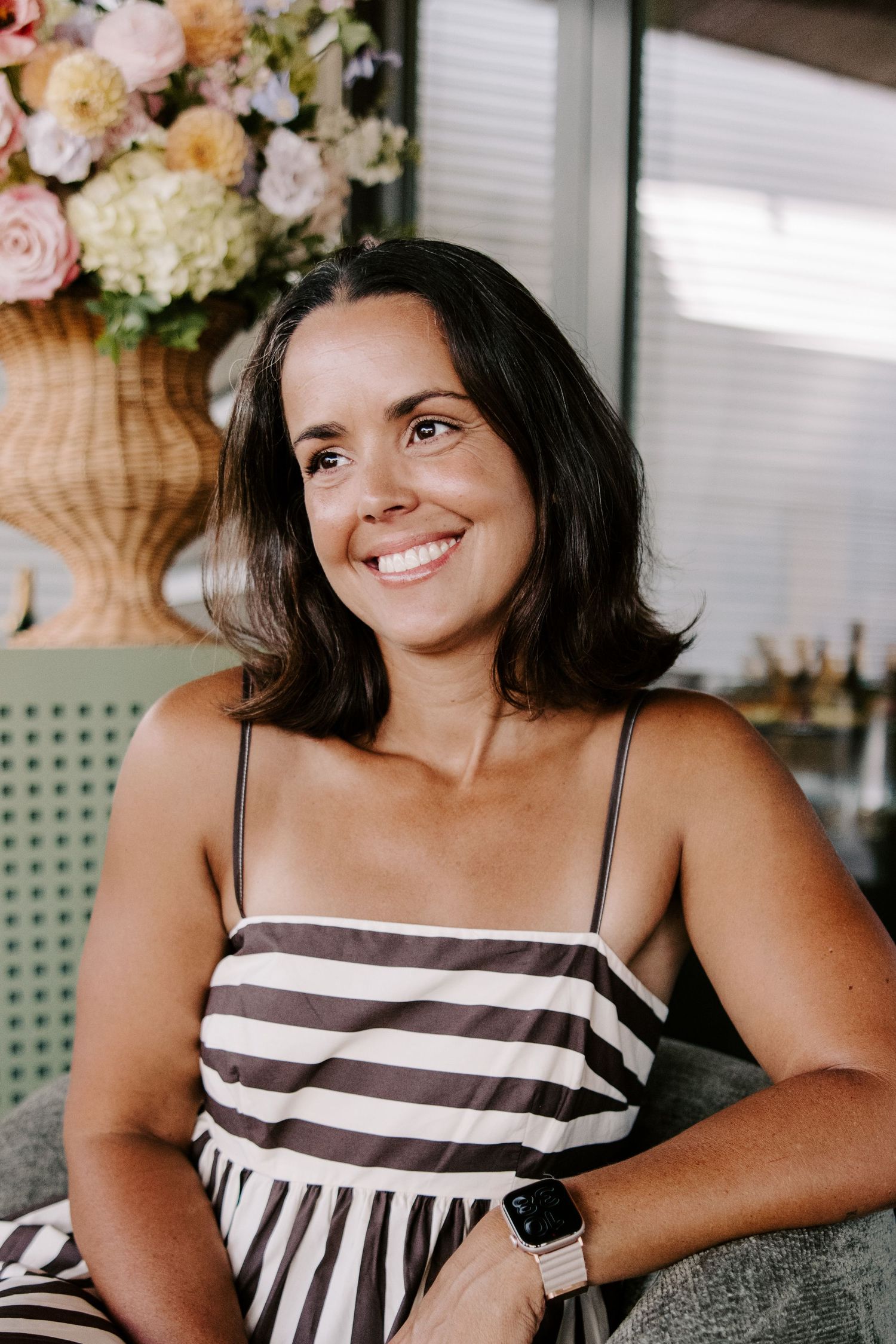 Smiling woman in striped sundress wearing Apple Watch sits relaxed indoors with floral arrangement in background.