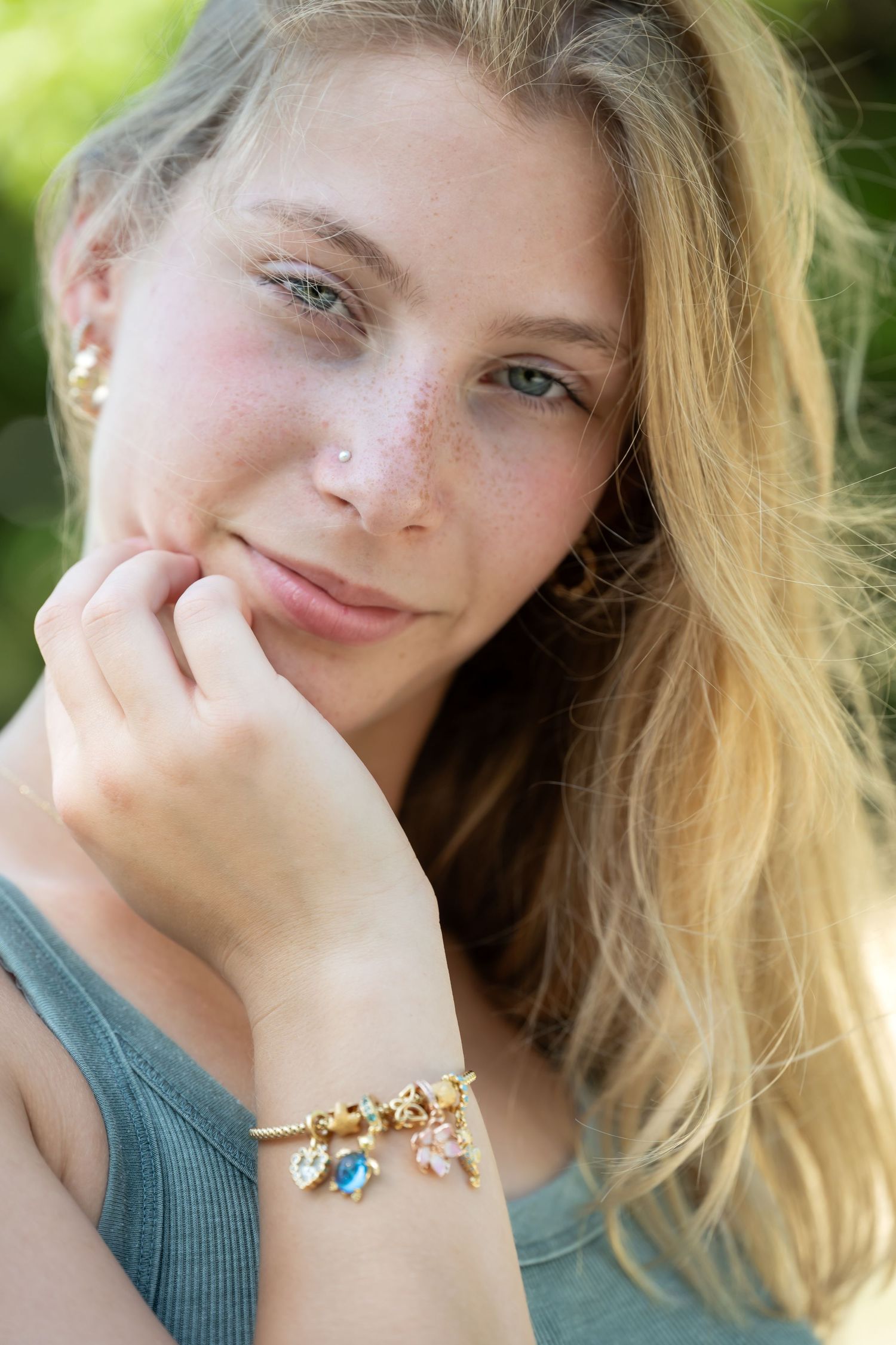 Young blonde woman with freckles wearing a charm bracelet, resting chin on hand outdoors.