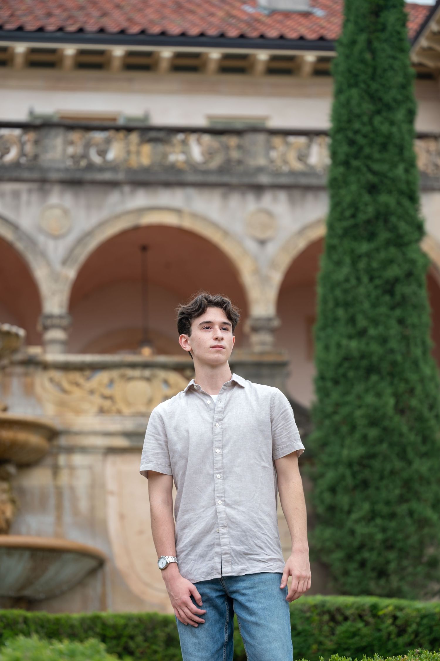 Young man in gray shirt and jeans stands in front of a historic building with arches and a fountain.
