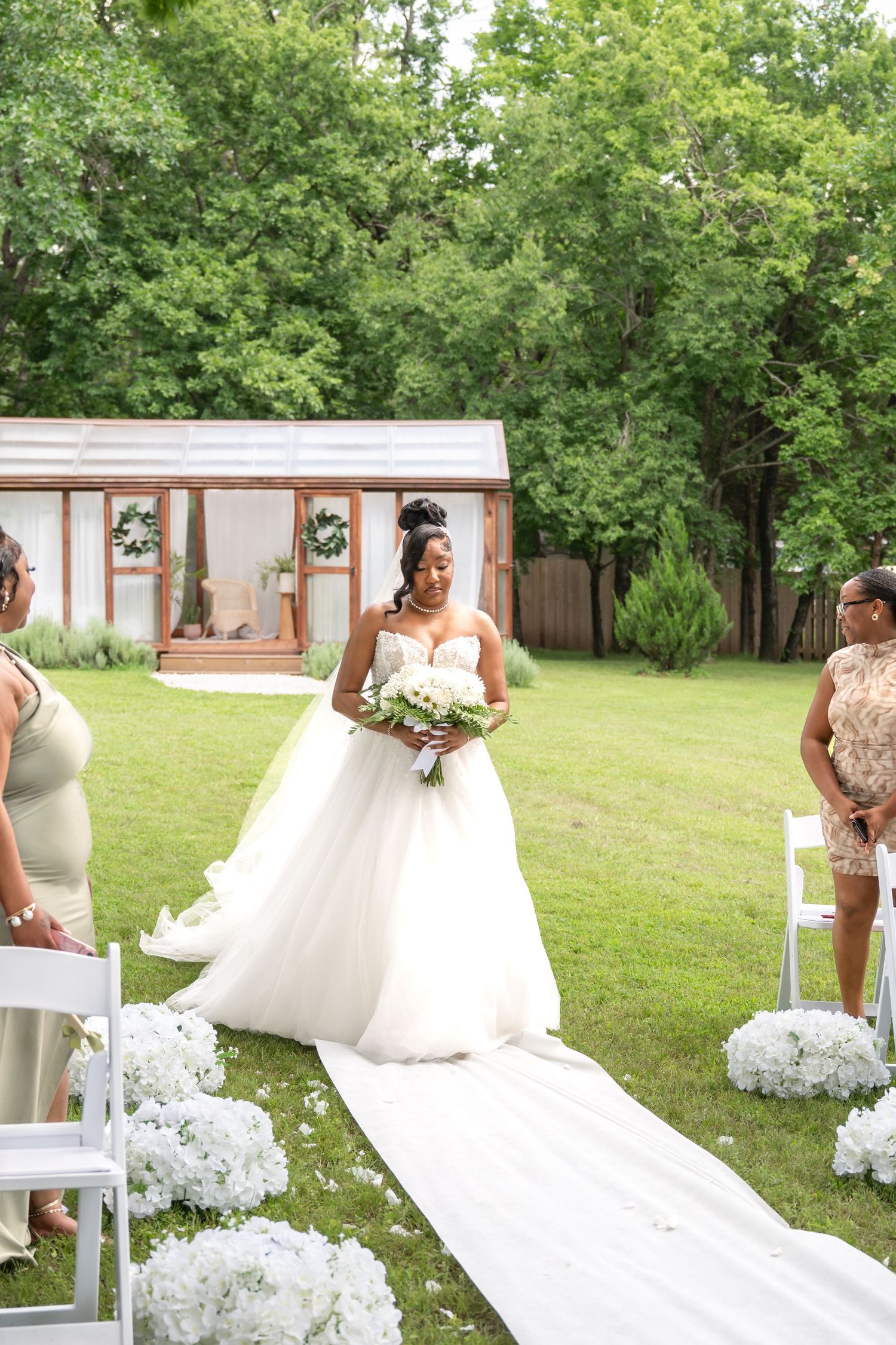 Bride in a white ball gown walks down an outdoor aisle lined with white florals toward a garden chapel.