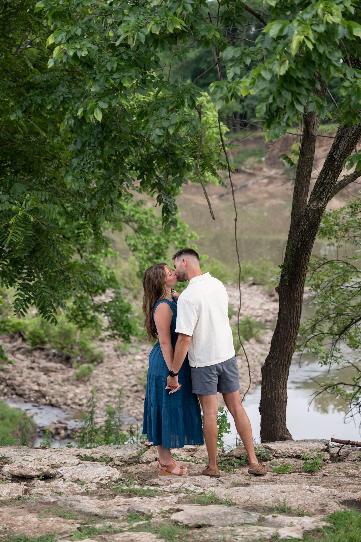Couple kissing under a tree by a rocky riverbank, woman in blue dress and man in white shirt and gray shorts.