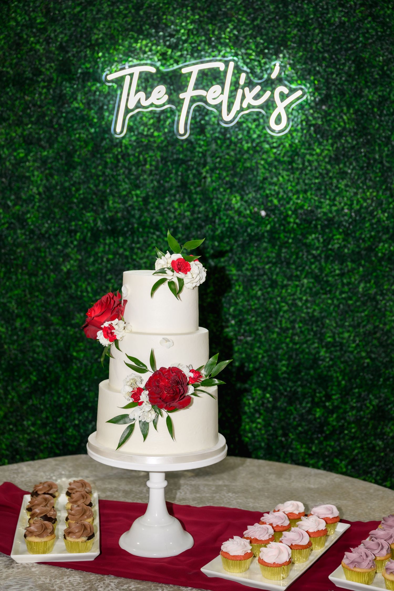 Three-tier white wedding cake with red floral decor under The Felix's neon sign, surrounded by cupcakes on a dessert table.