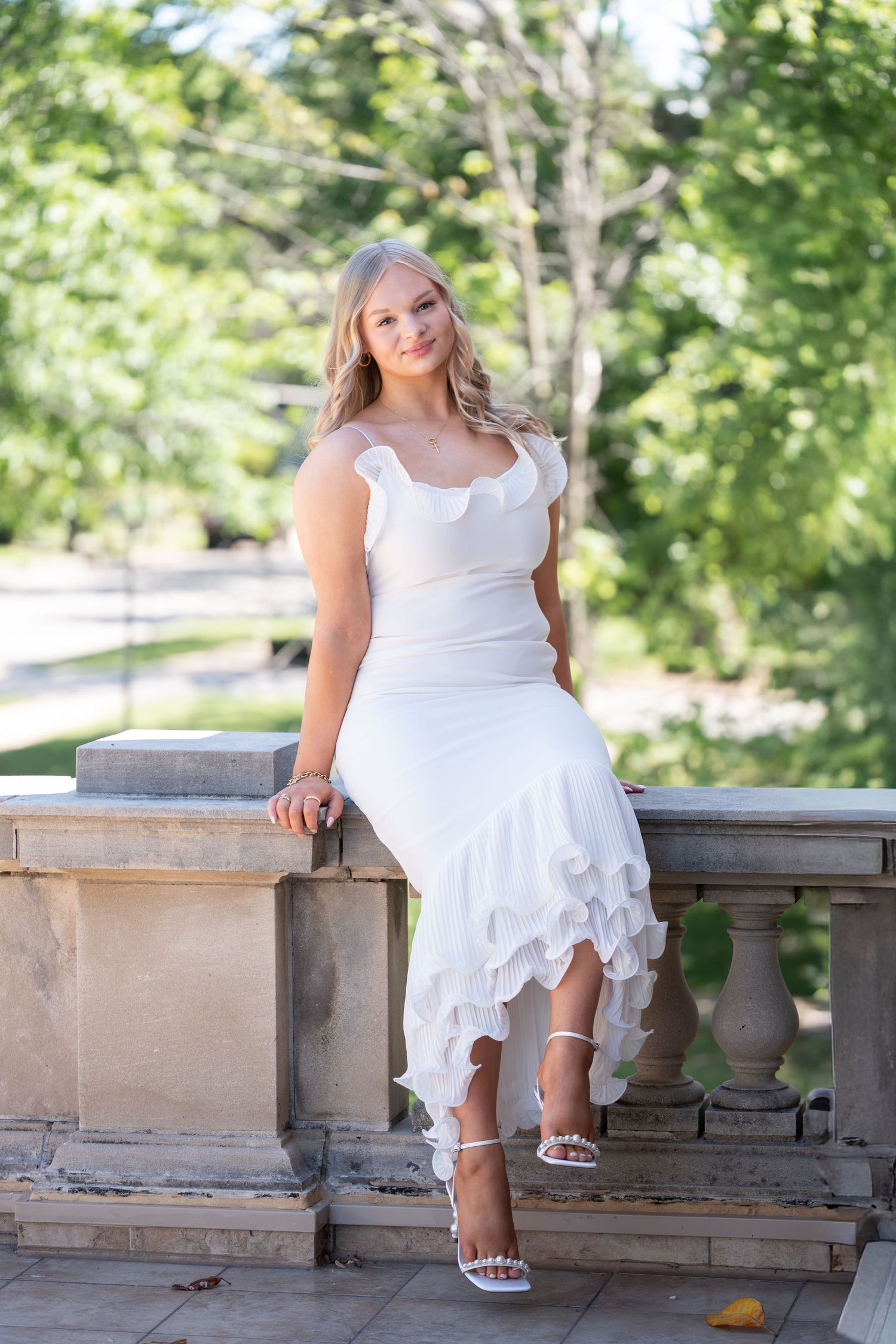Young blonde woman in white ruffled dress and heels poses on stone balustrade in sunny garden setting.