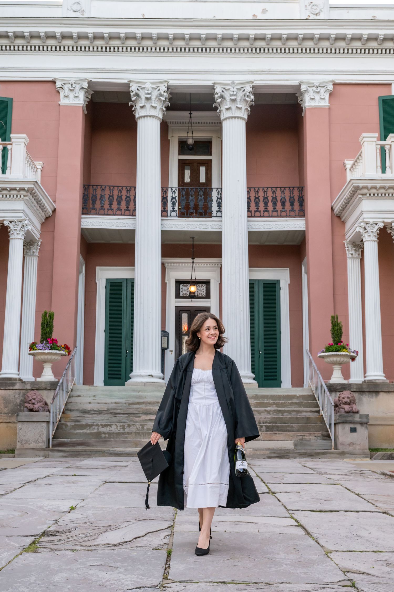 Woman in graduation gown and cap walks in front of a grand pink colonial building with white columns.