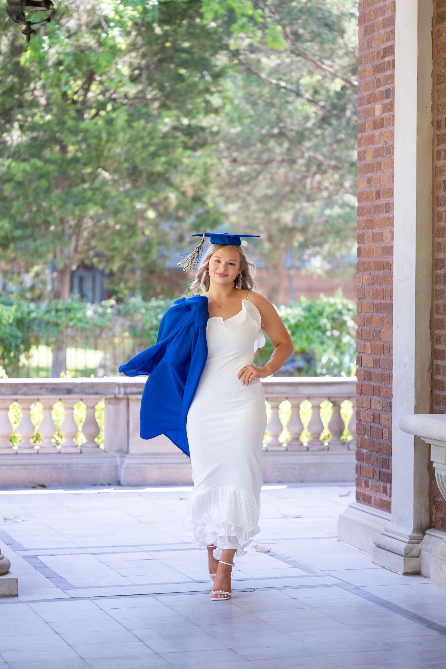 Young woman in white dress and blue graduation cap and gown walks confidently on a college campus colonnade.