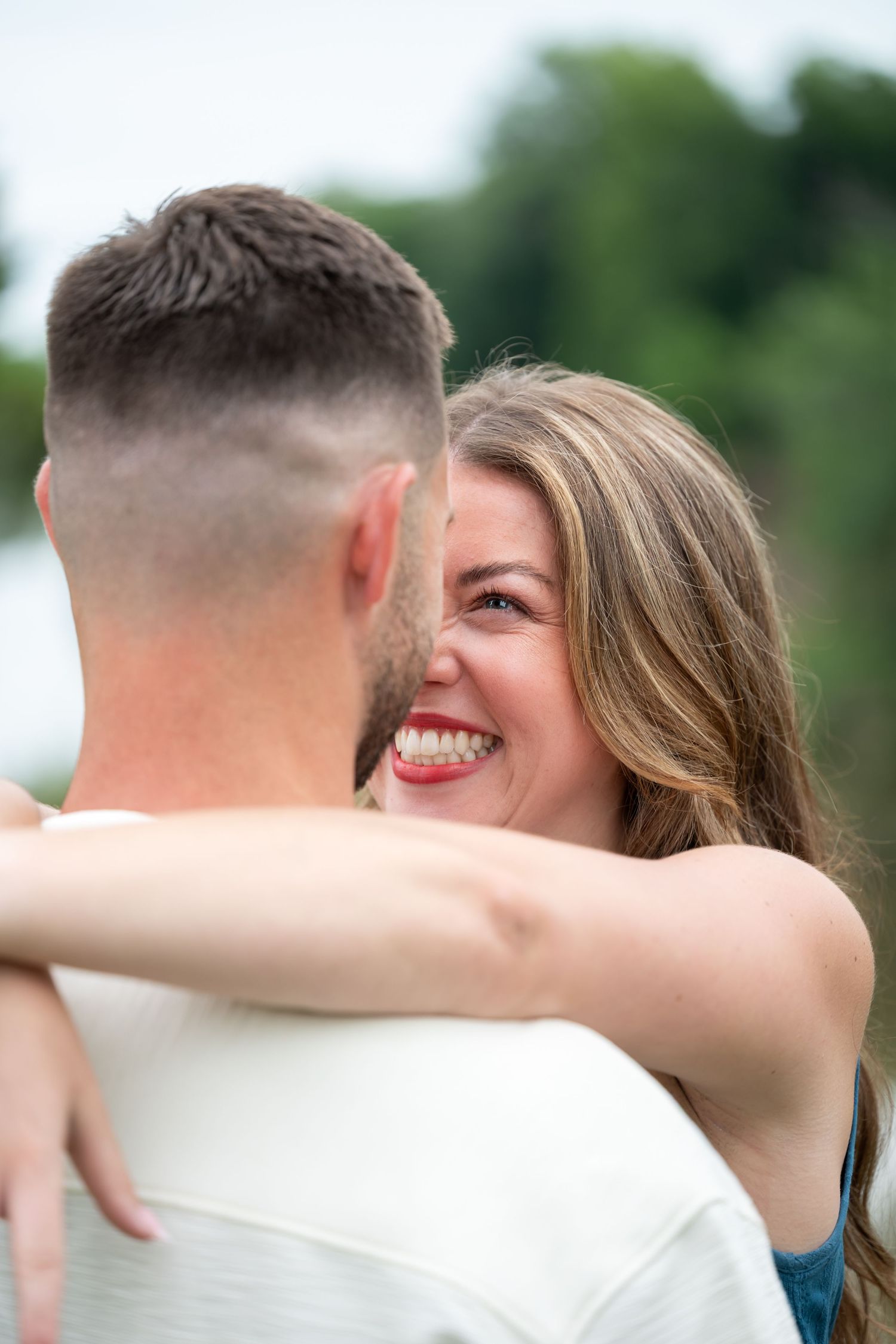 A smiling woman with long hair embraces a man outdoors, looking joyfully over his shoulder near greenery.