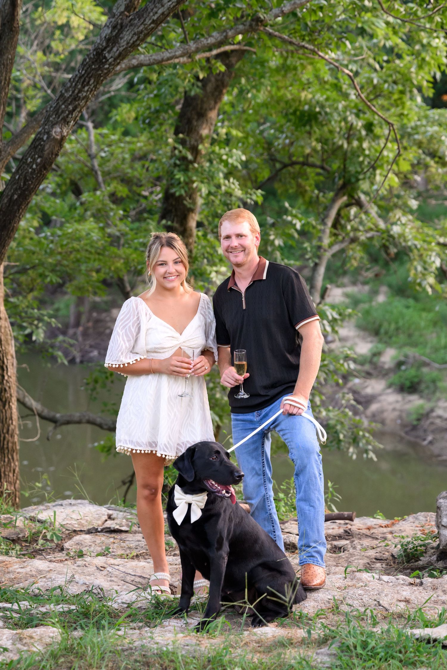Couple holding champagne glasses with black Labrador dog on leash, standing by a creek surrounded by lush green trees.