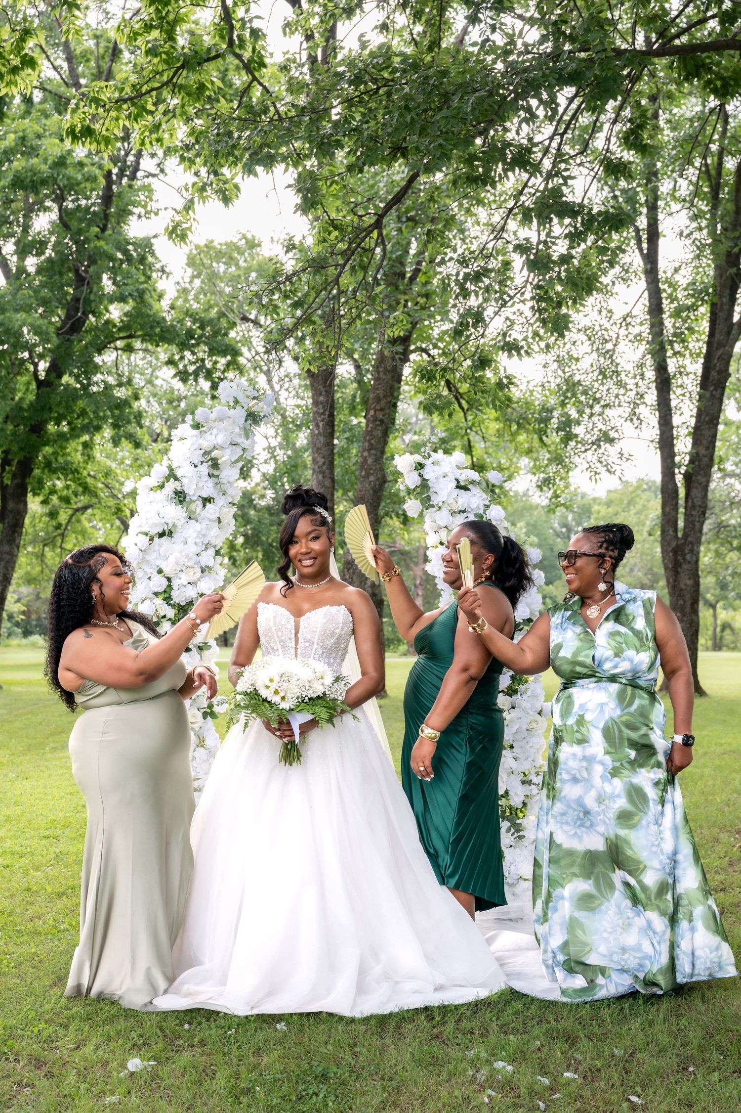 Bride in white gown poses with three bridesmaids in green dresses near floral arch outdoors.