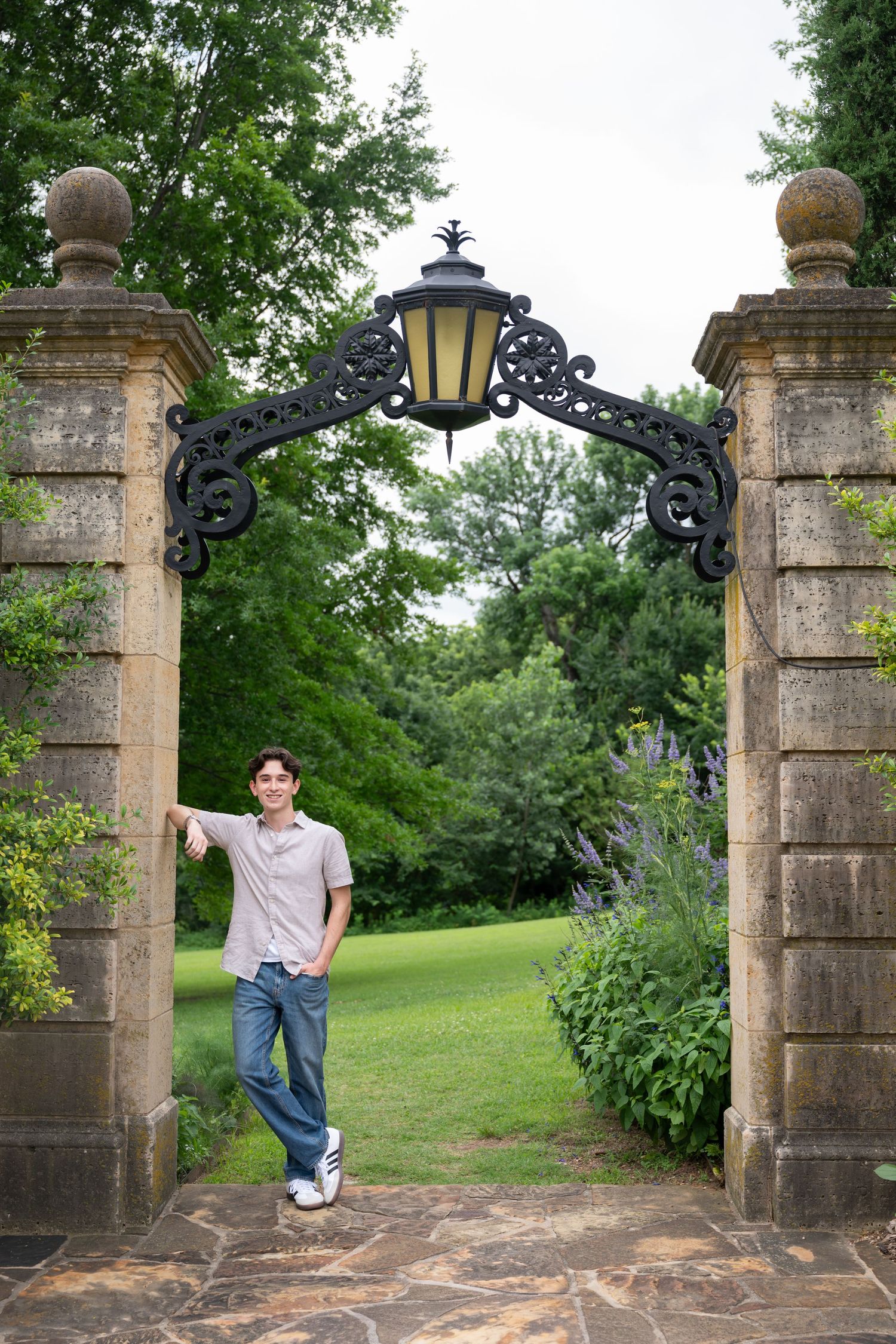 Young man leaning against decorative iron gate with lantern between stone pillars in a lush green garden.