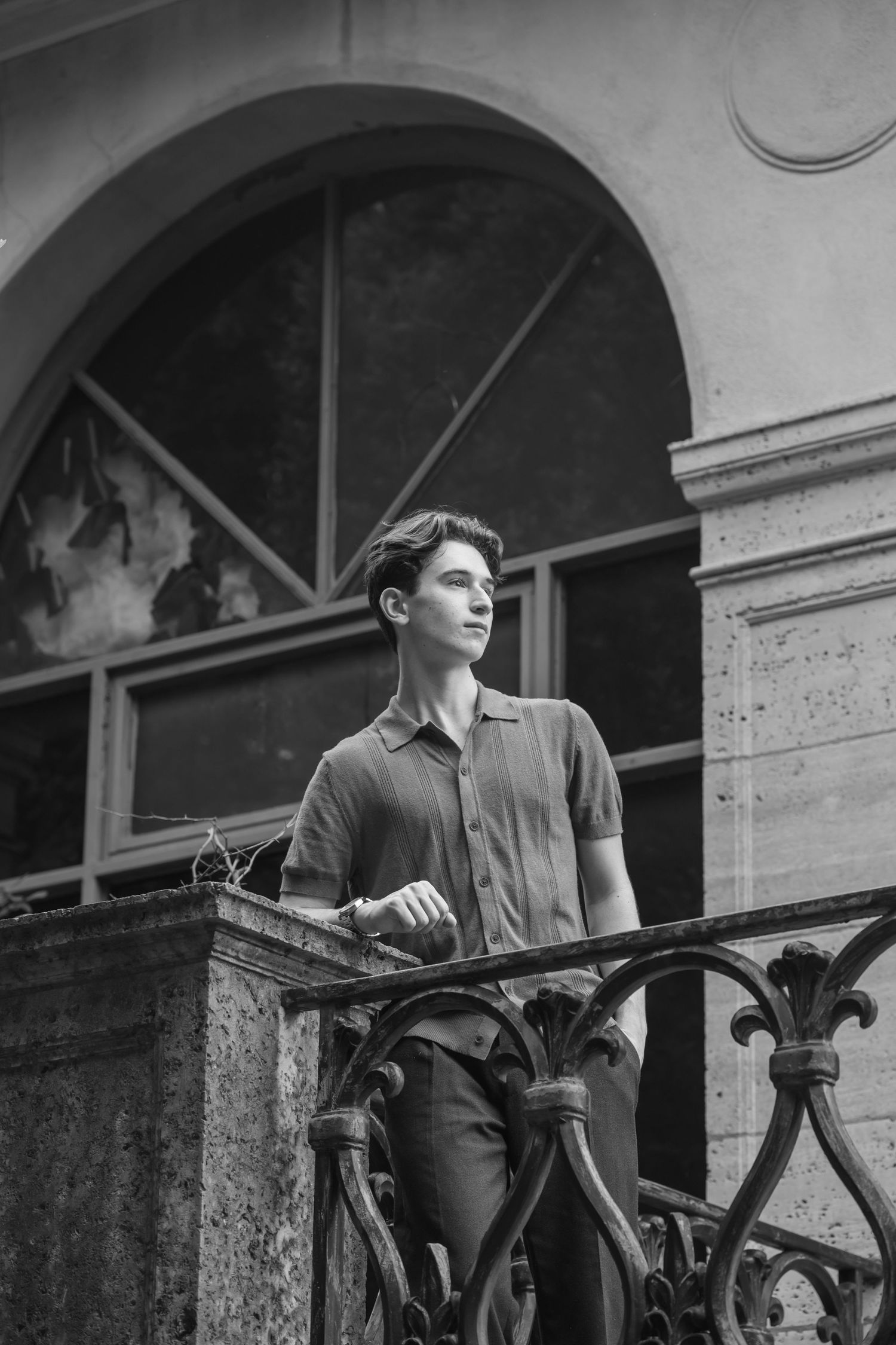 Young man in polo shirt stands on ornate iron balcony of historic stone building in black and white photo.