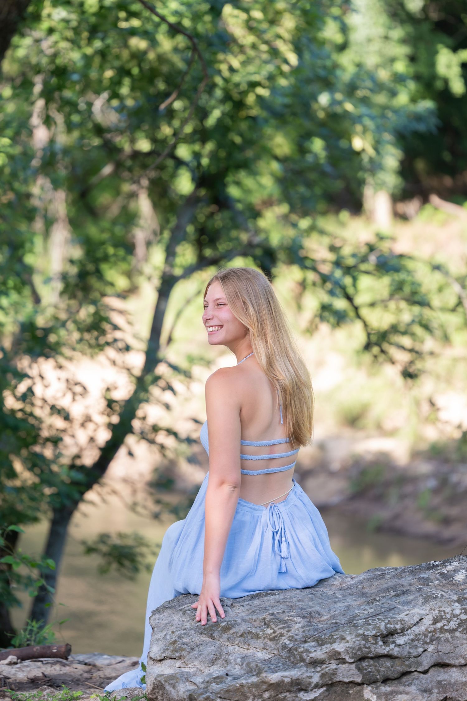Smiling blonde woman in blue strappy crop top and skirt sitting on rocks near a forest creek.