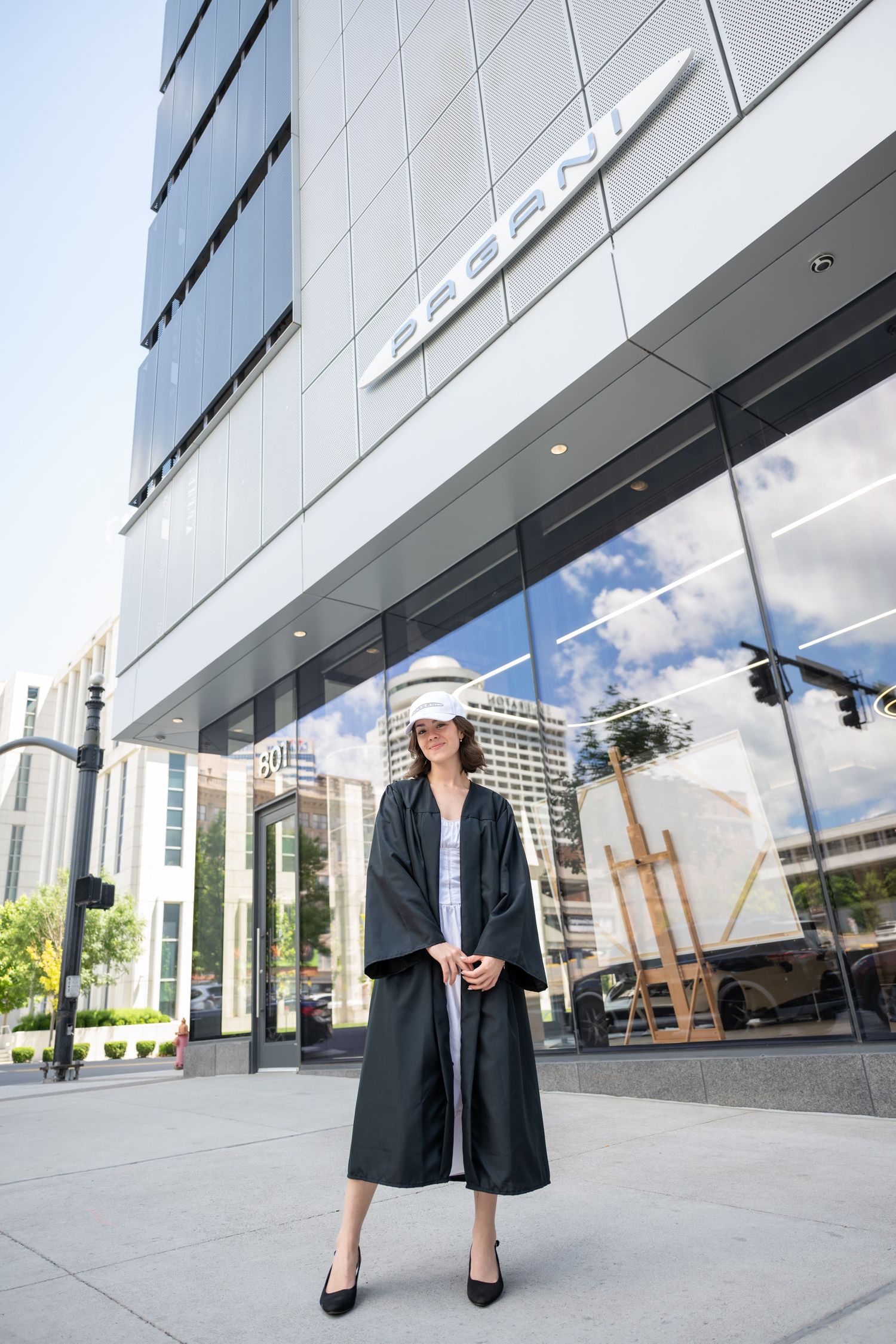 Graduate in black gown and white hat stands confidently outside a modern glass building on a sunny day.