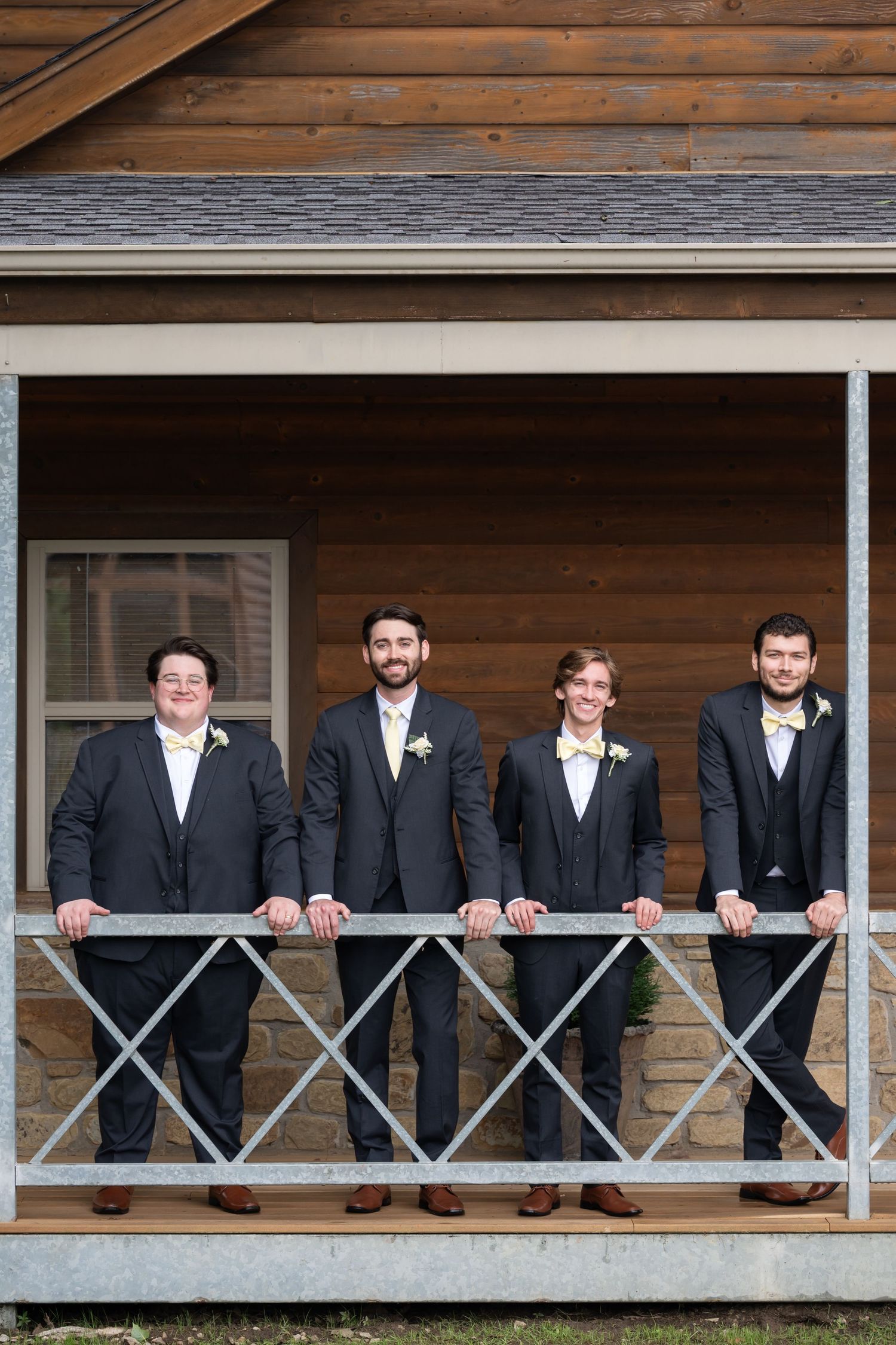 Four groomsmen in dark suits with yellow bow ties pose on a rustic wooden cabin porch for a wedding photo.