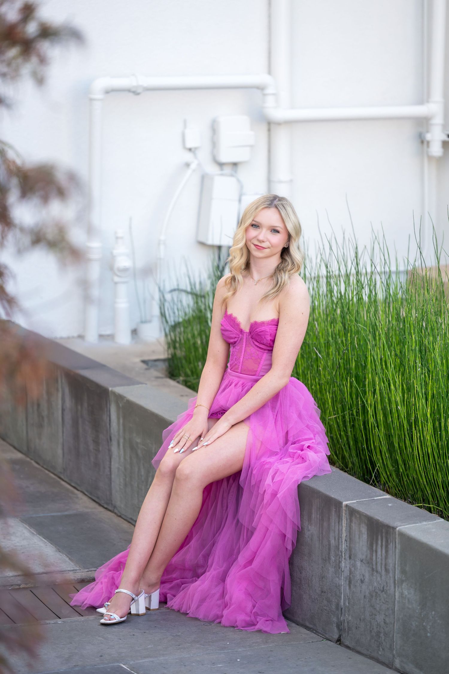 Young woman in a strapless pink tulle gown sitting on a stone ledge outdoors with green grass behind her.