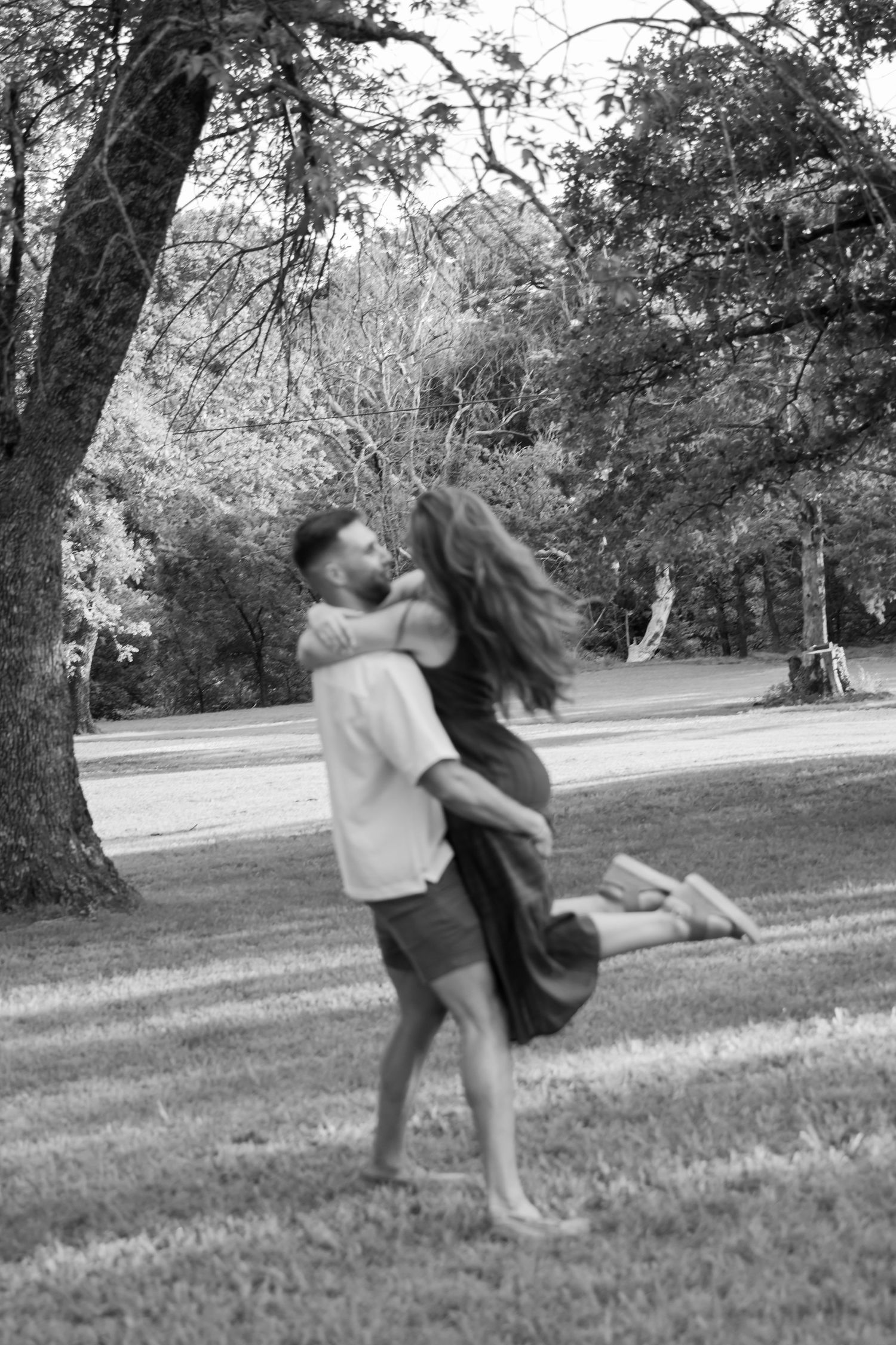 Black and white photo of a man lifting and spinning a woman in a park, surrounded by trees.