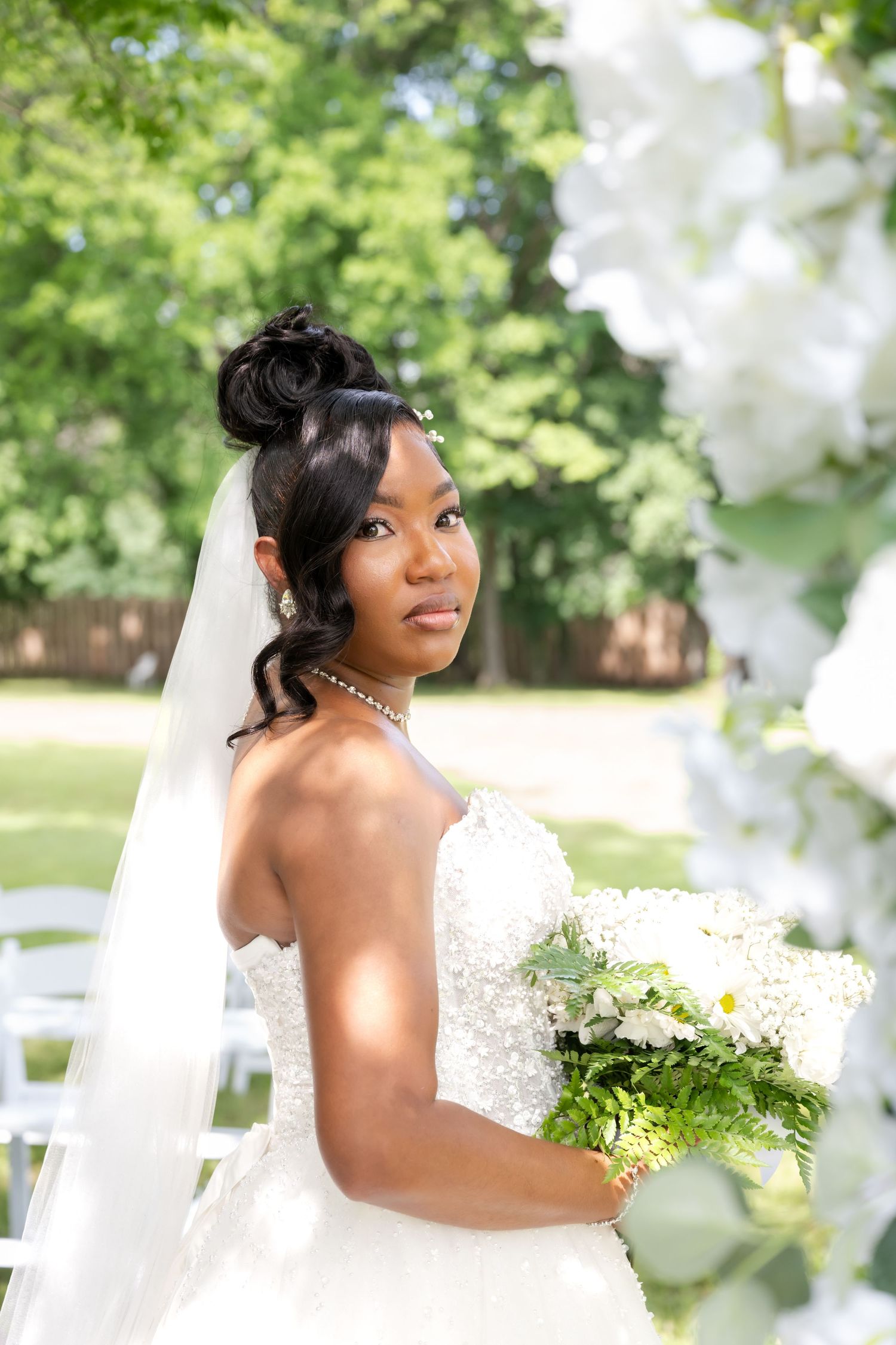 Elegant Black bride in white strapless gown holding bouquet, wearing veil and pearl necklace at outdoor wedding.