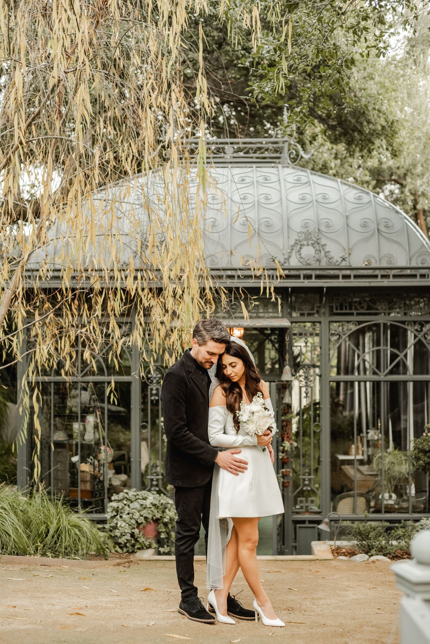 Elegant wedding photos near a greenhouse with willow trees creating natural framing.