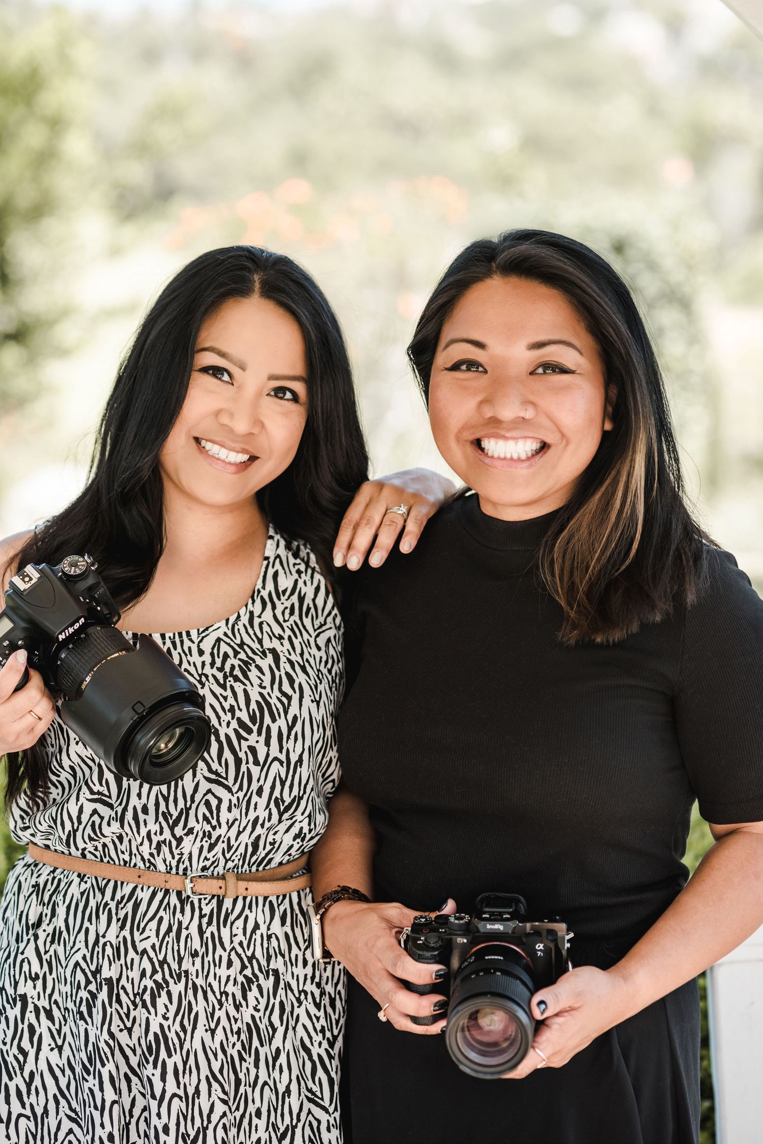 Two smiling women photographers holding DSLR cameras outdoors, wearing stylish outfits in a bright natural setting.