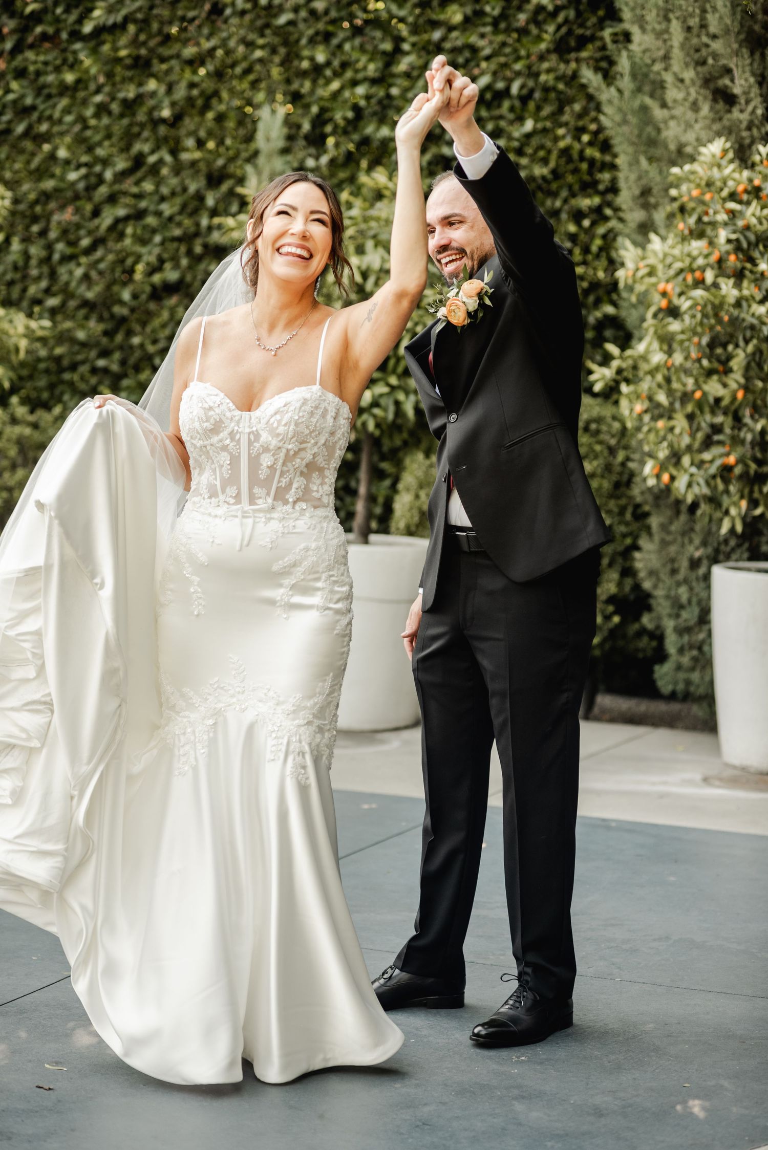 A bride in a fitted lace gown and groom in black tuxedo dance joyfully on an outdoor patio with ivy-covered walls.