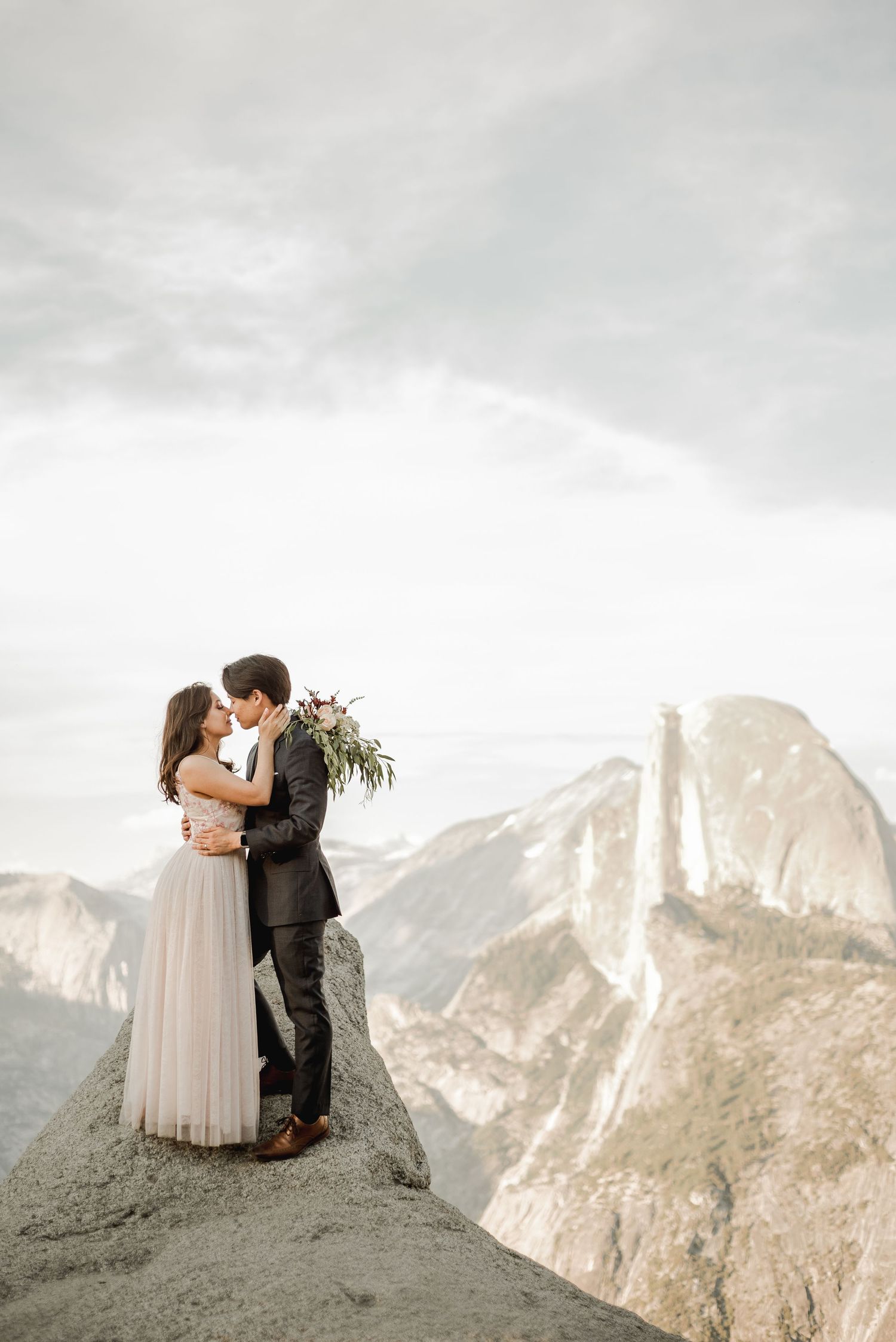 Couple sharing romantic moment on mountain cliff with dramatic valley vista behind them.
