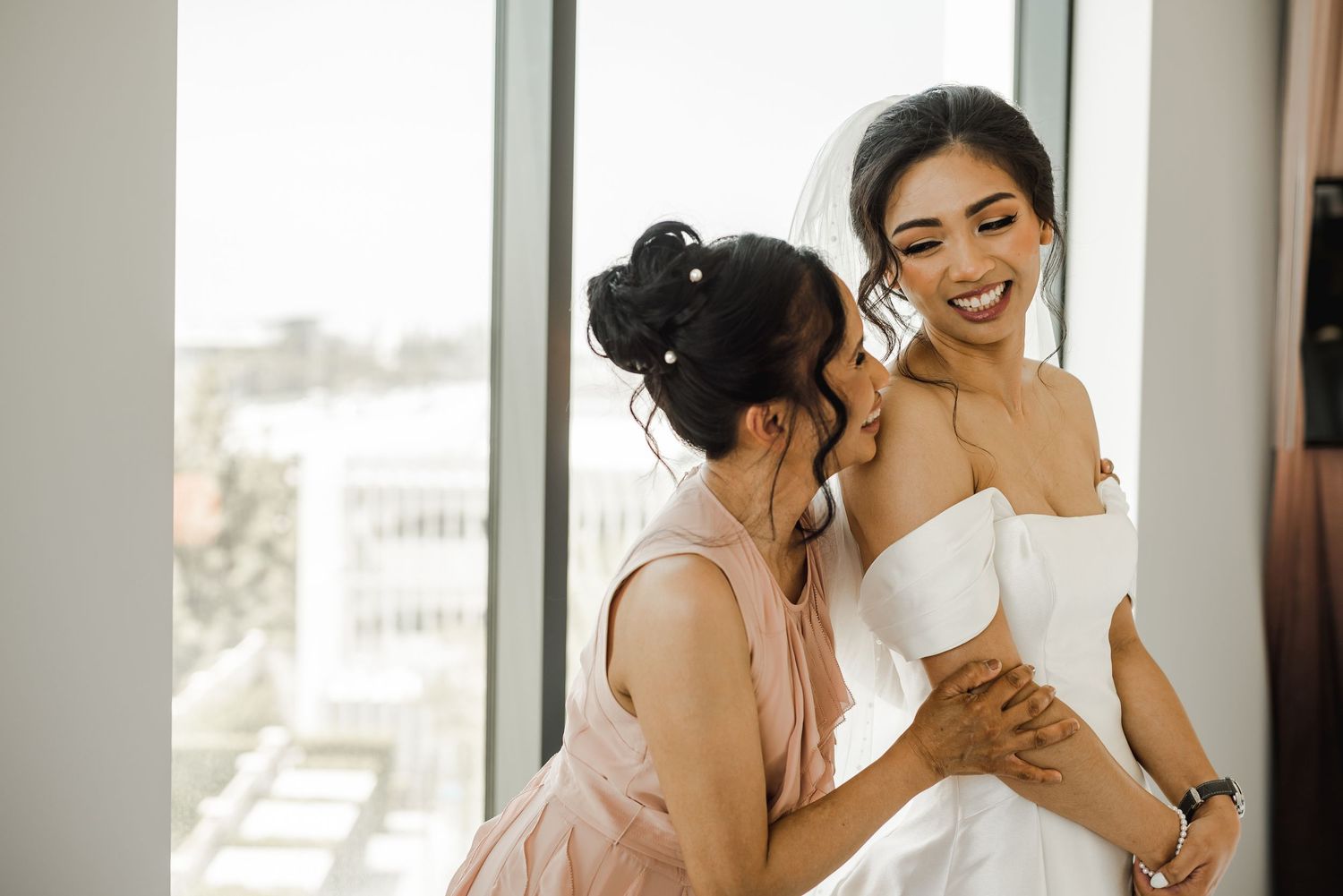 Two women in white dresses share joyful moments together during a wedding preparation.