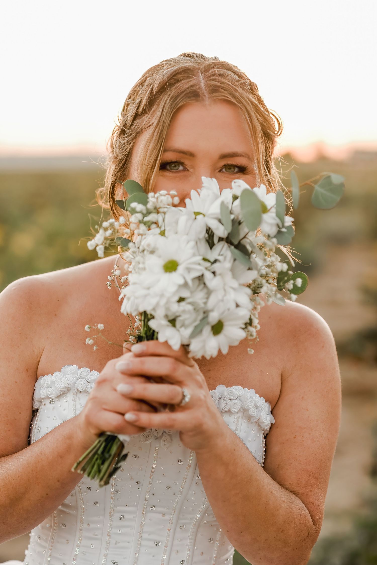 A bridal portrait showing a white daisy bouquet with eucalyptus held up against a sunset background.