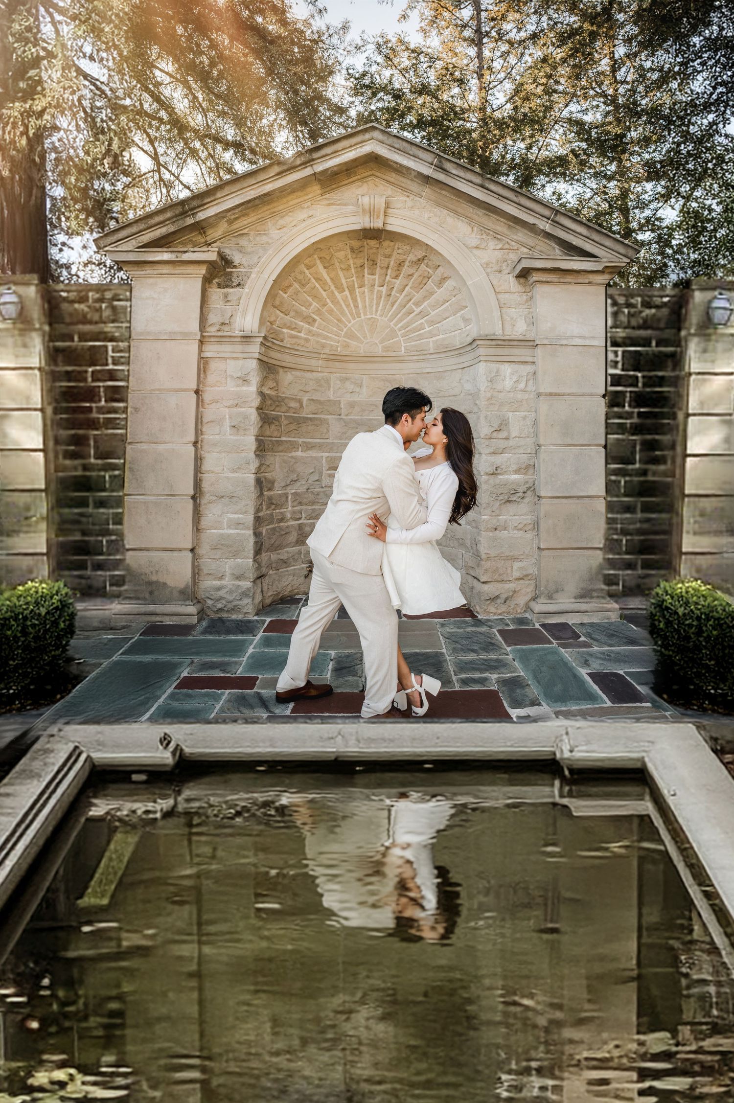 Couple in white outfits creates artistic reflection poses by ornate stone fountain.