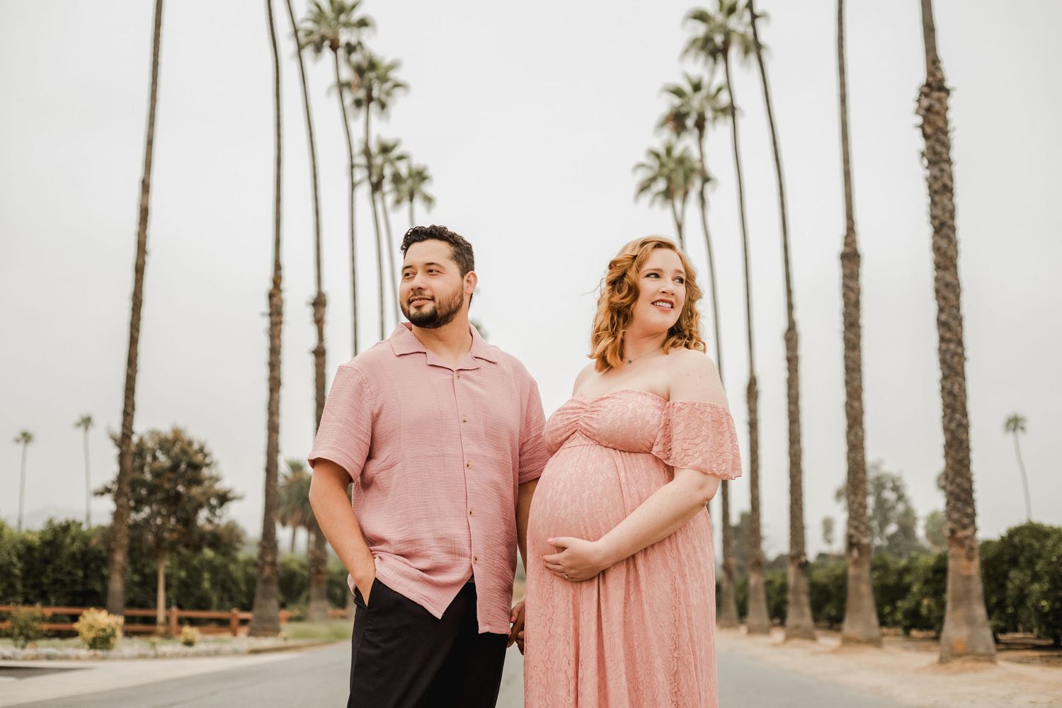 A couple poses for maternity photos in coordinating pink outfits with palm trees in the background.