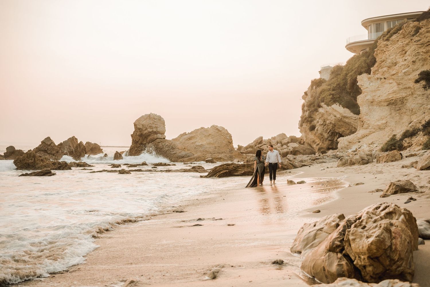 Couple walking along misty beach coastline with rocky formations.