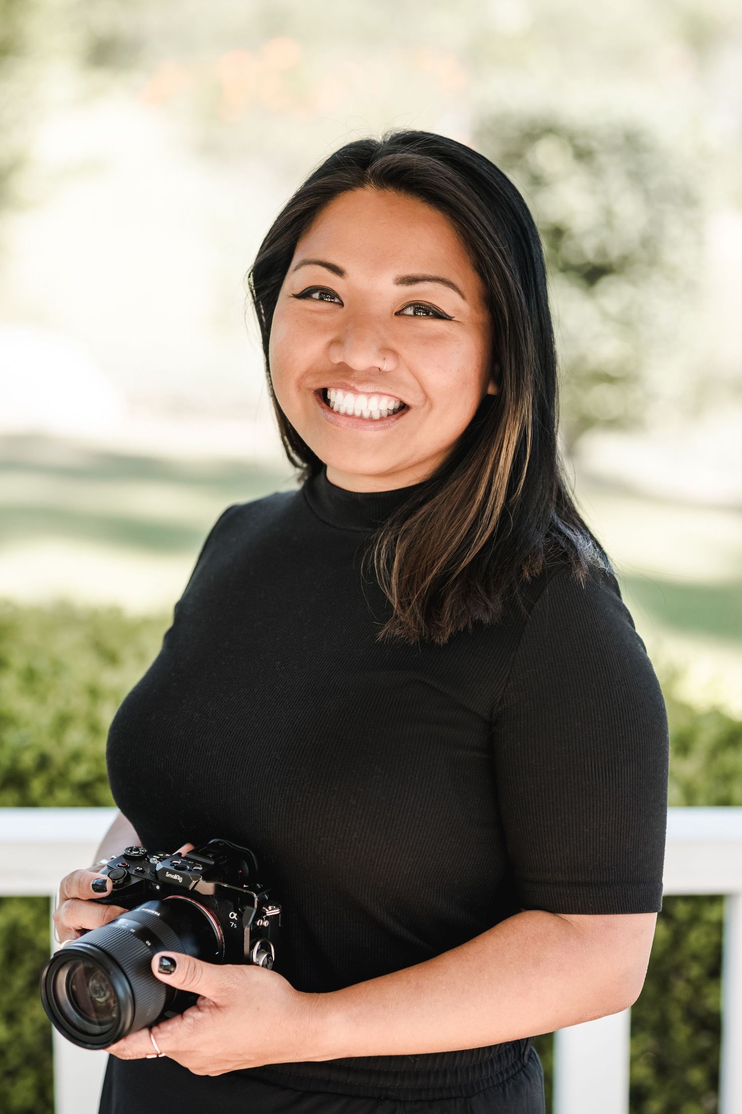 Professional female photographer in a black top smiles while holding a camera outdoors in natural lighting.