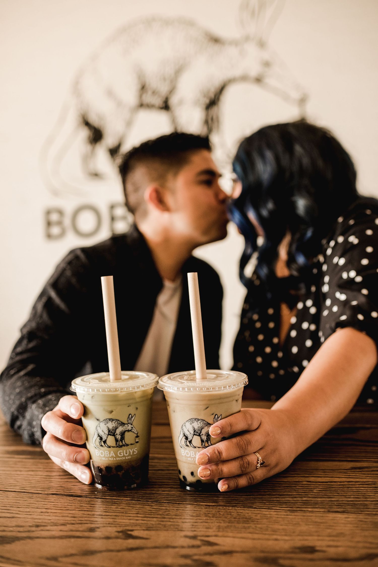 Couple sharing boba tea drinks at wooden table in cafe setting.