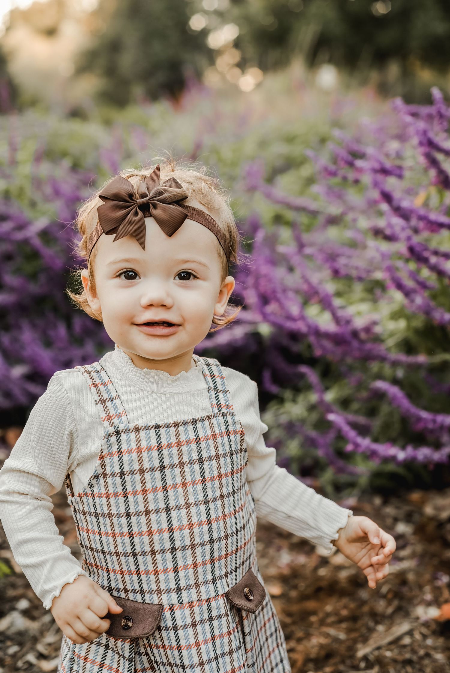 A toddler in plaid overalls and bow headband smiles among purple flowers.
