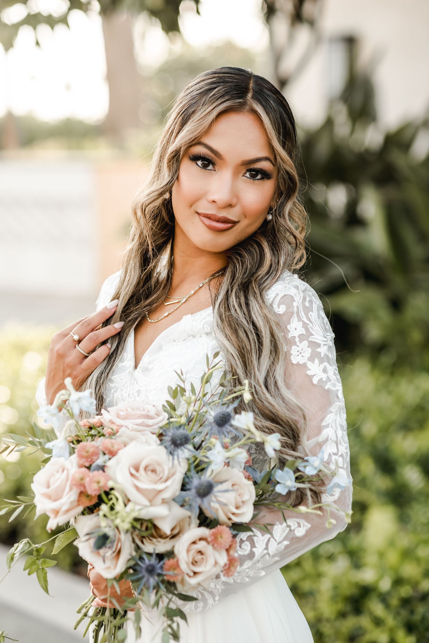 Elegant bride with long wavy hair holding pastel bouquet against garden backdrop.