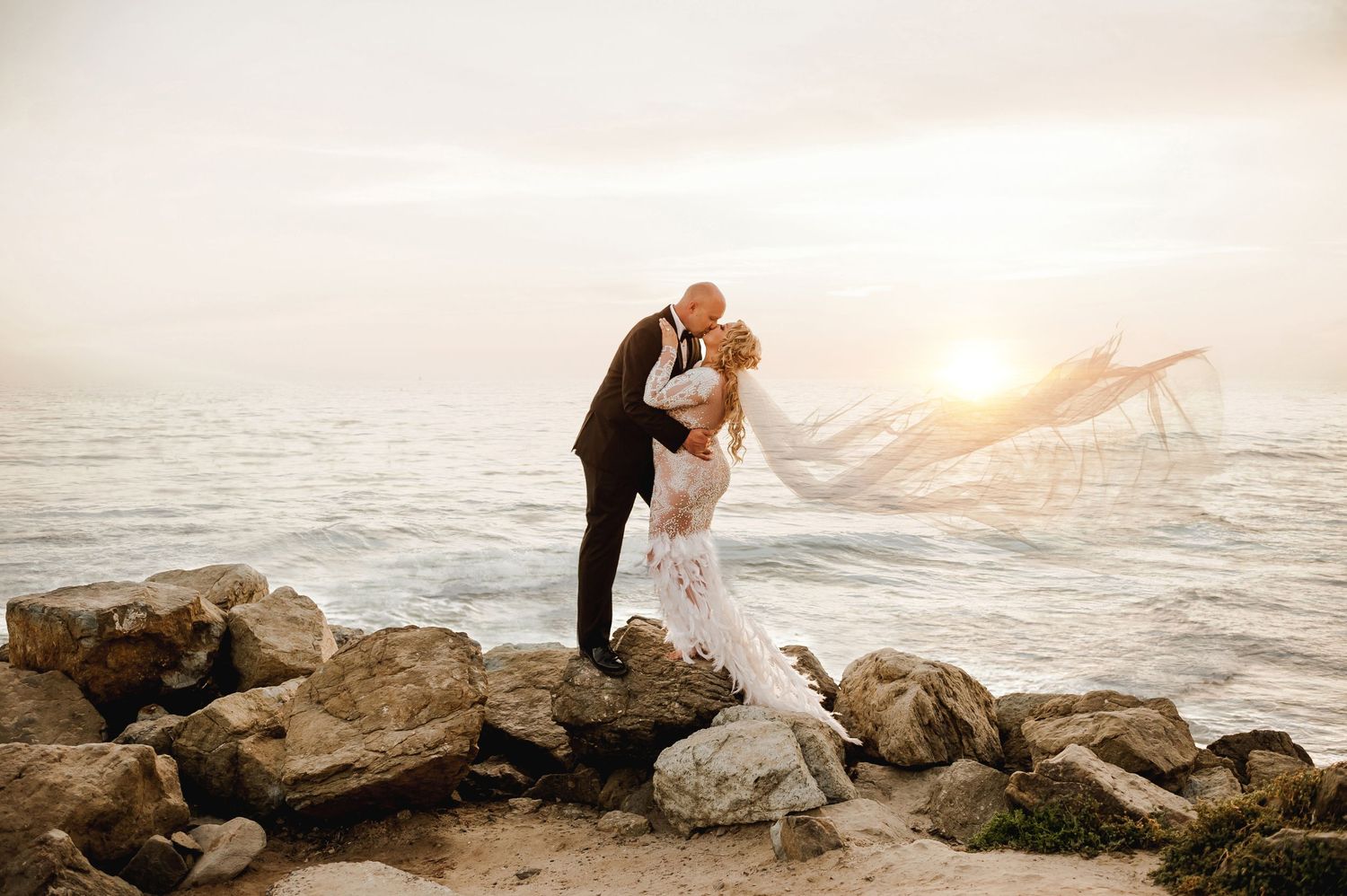 A romantic sunset wedding photo with a couple embracing on rocky coastal shores.
