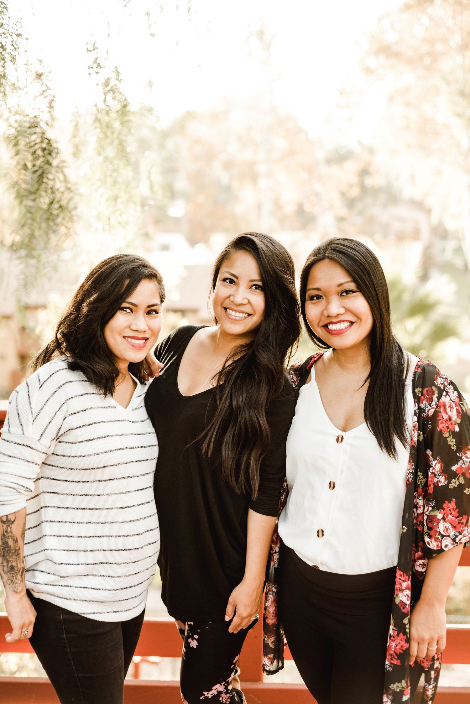 Three young women in casual attire smiling together outdoors on a sunny day.