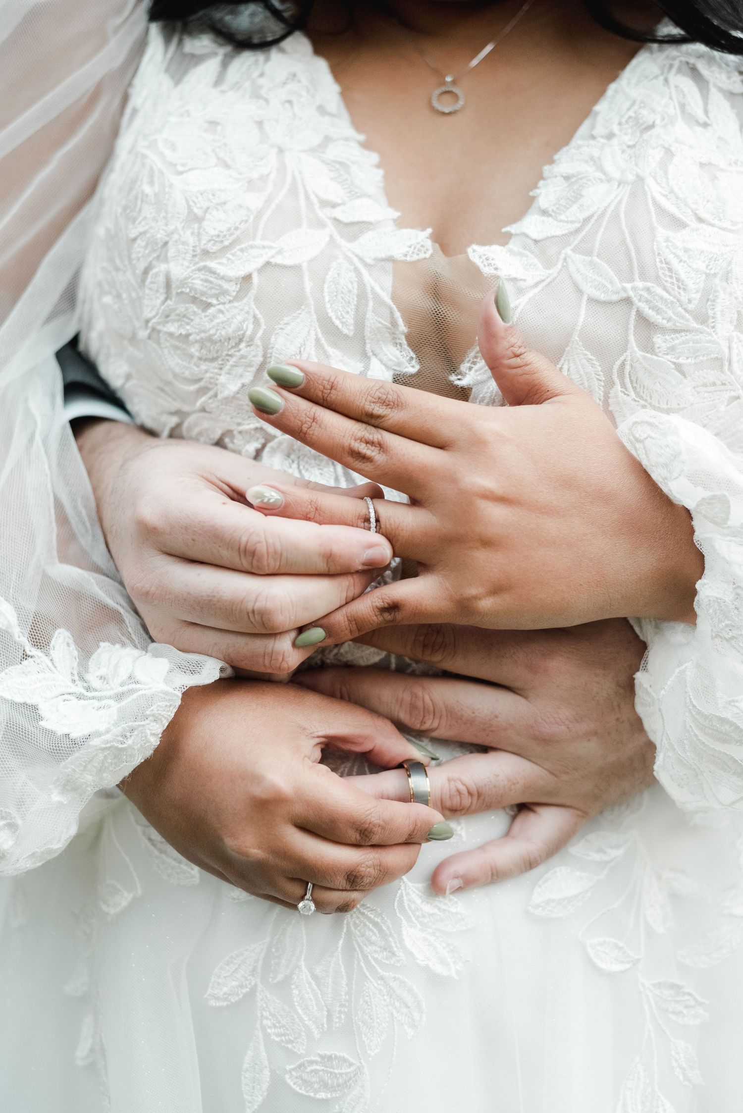 Close-up of hands with wedding rings against delicate white lace dress details.