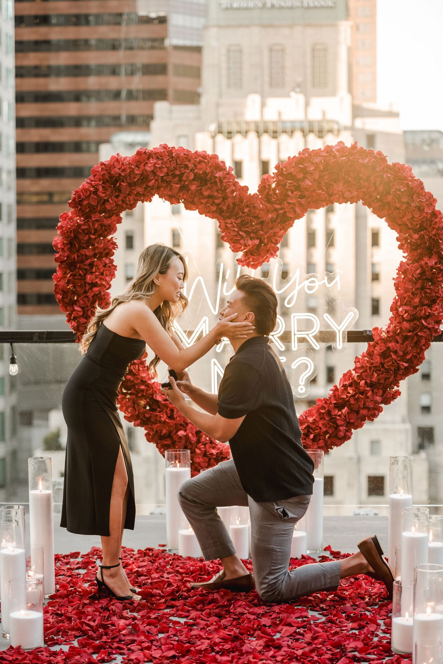 A proposal scene with a large red heart arch and rose petals on a city rooftop.