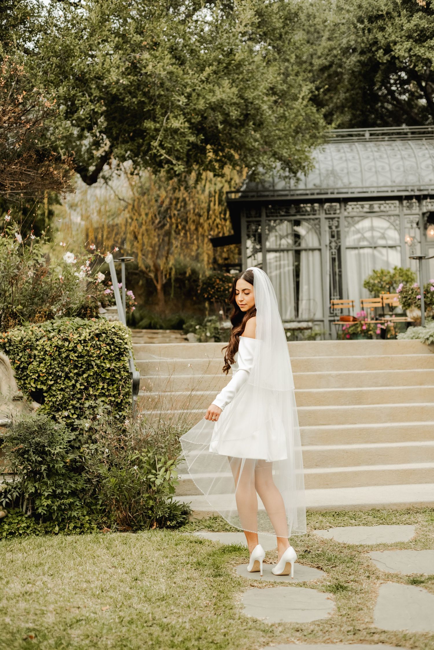 A bride in a short white dress poses on garden steps near a greenhouse.