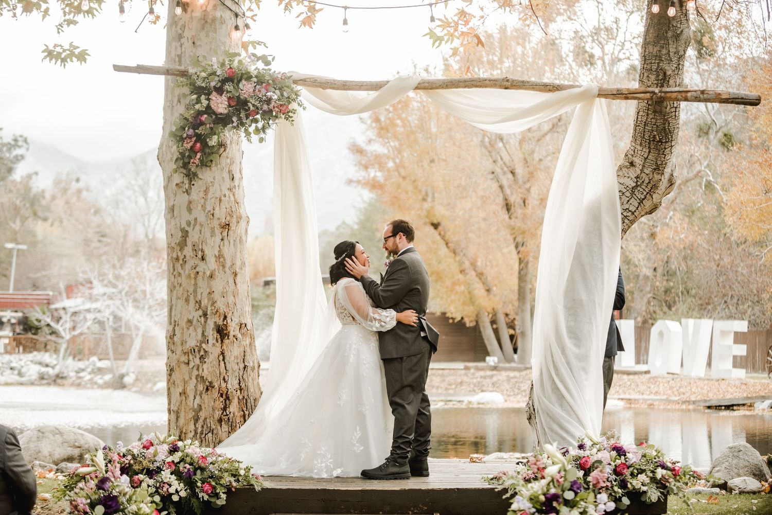 A couple shares a romantic moment under a floral arch with flowing white fabric.