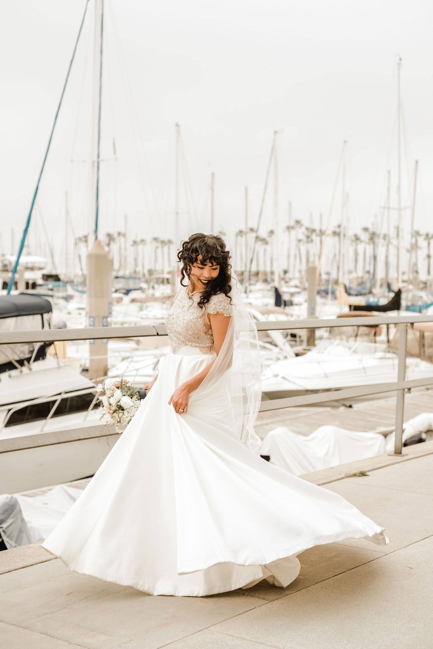 A bride twirling in a flowing white dress at a marina with sailboats in the background.