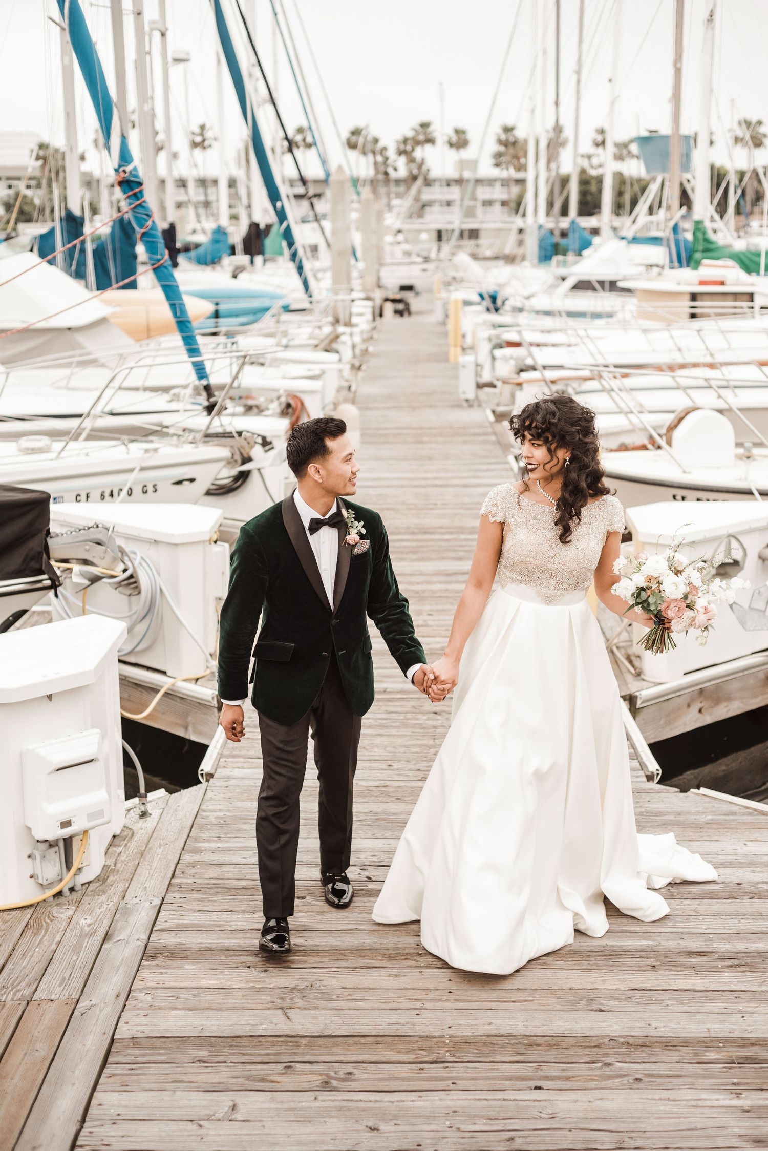 A couple walks hand in hand along a marina dock surrounded by sailboats.