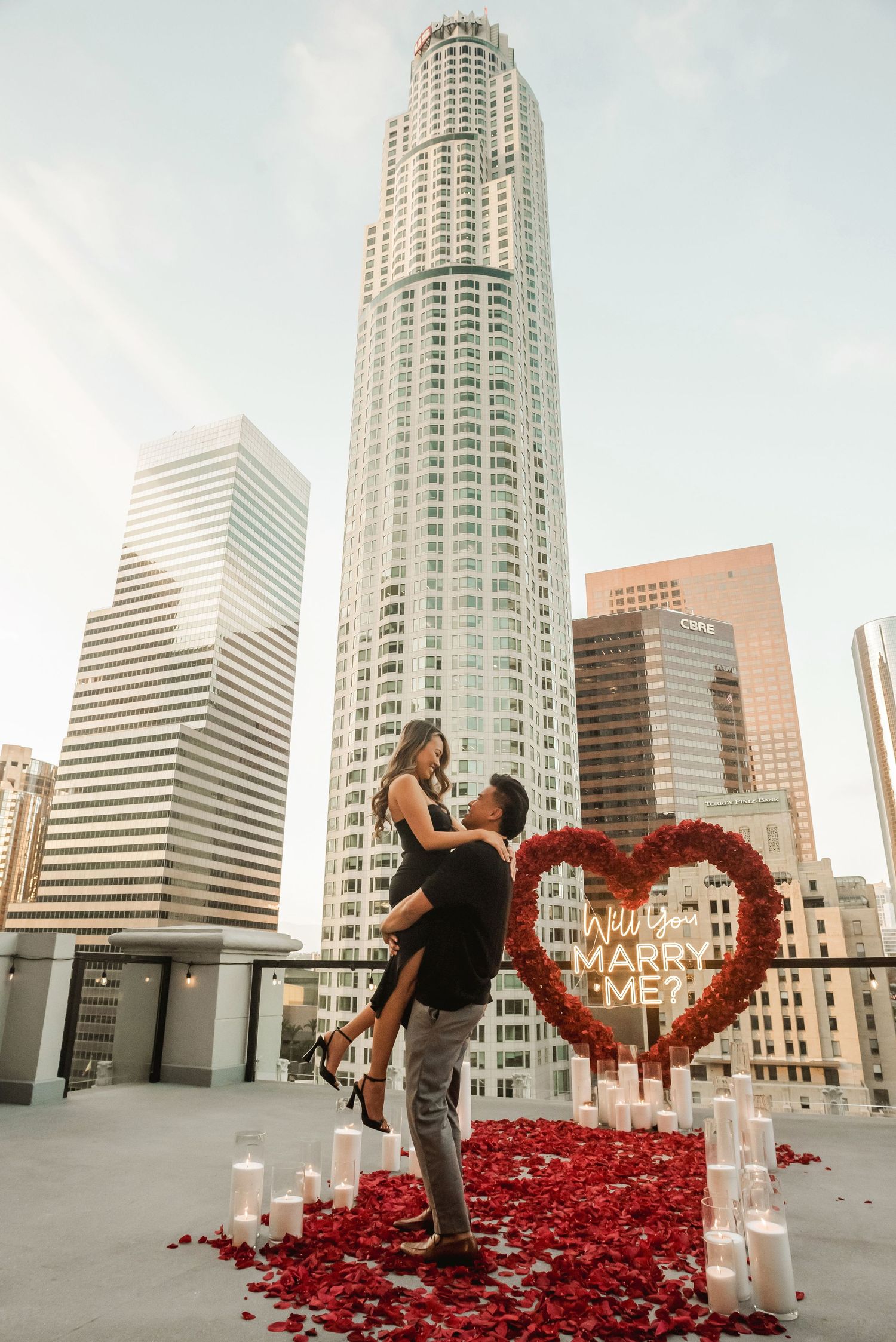 A romantic rooftop proposal in downtown Los Angeles with a red heart arch and city skyline.